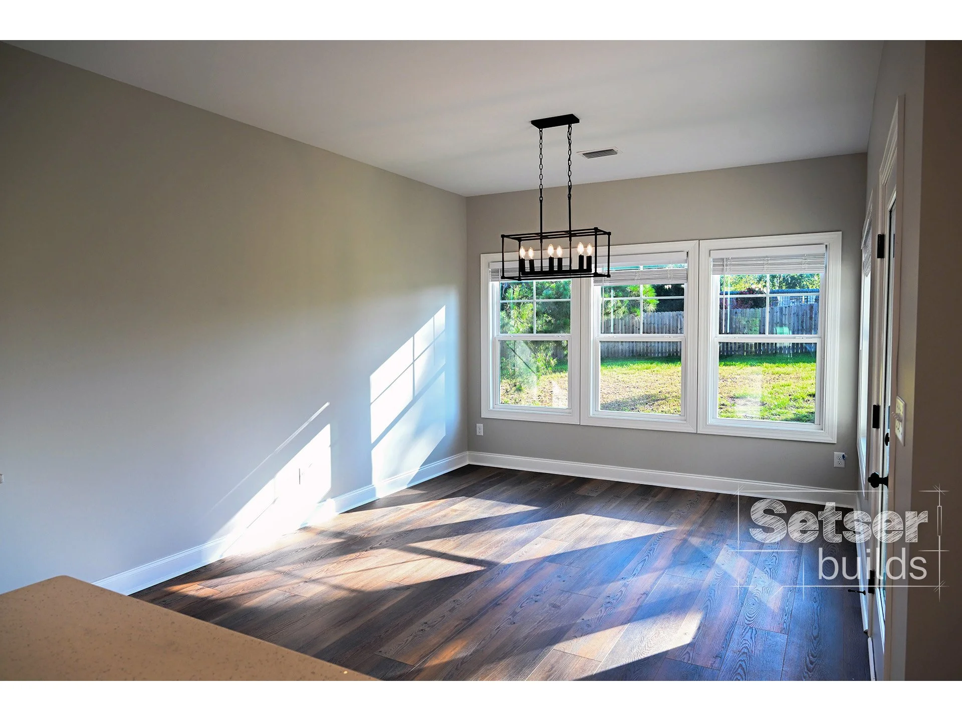 Bright empty room with hardwood flooring, large windows, and a modern black chandelier.