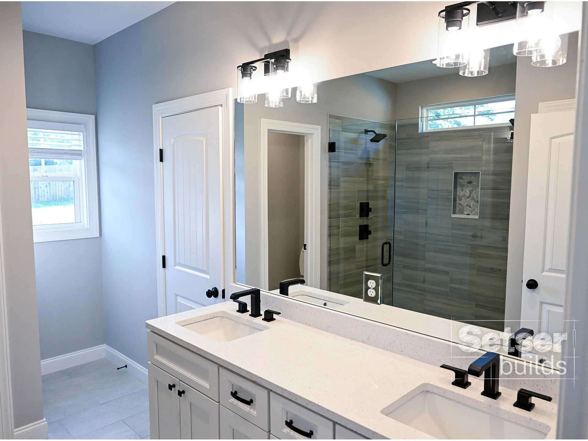 Bathroom with double vanity, black faucets, large mirror, walk-in shower with glass door, grey tile walls, small window, white door, and light grey walls.