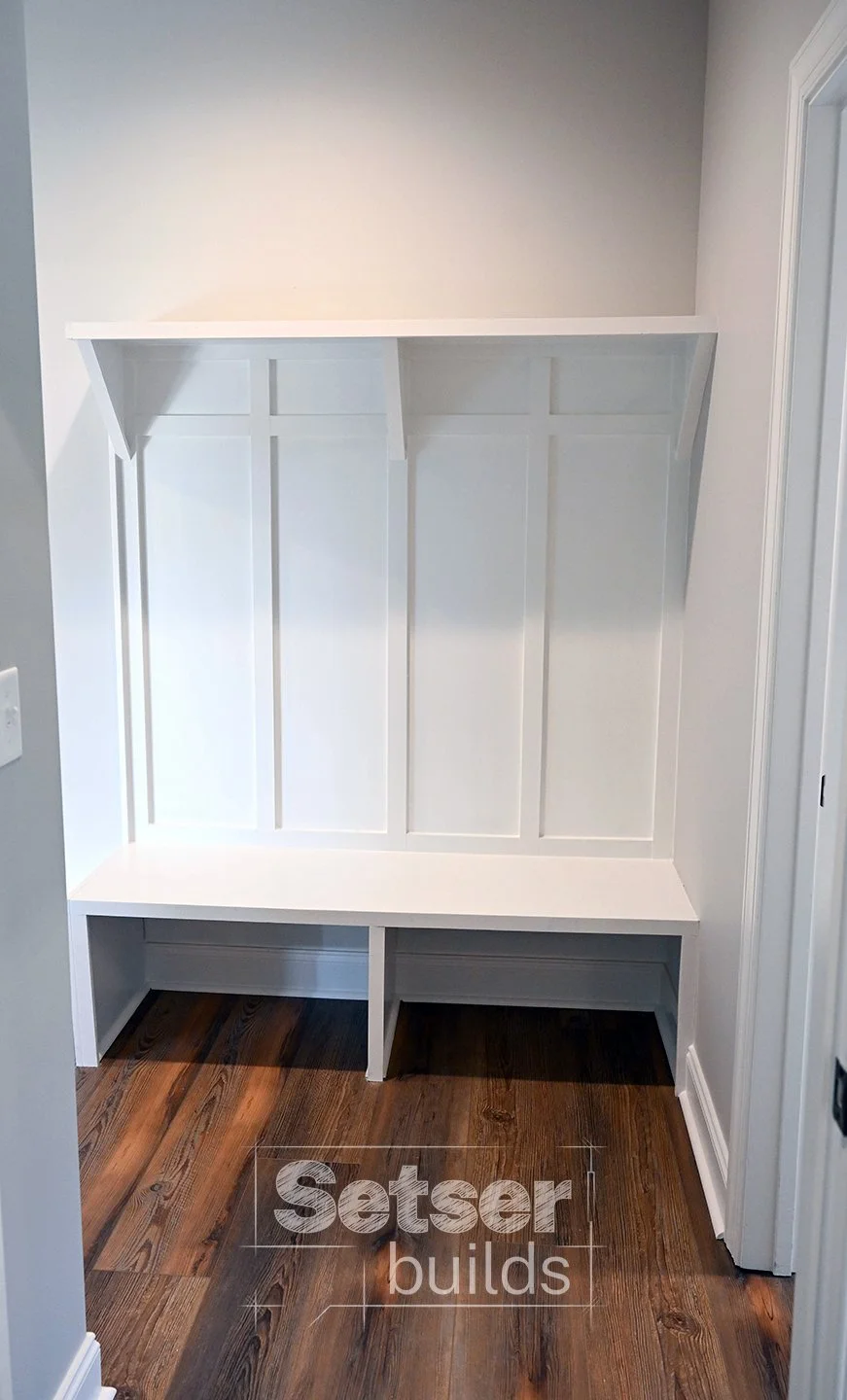 White built-in mudroom or entryway bench with storage space underneath and a shelf above, against a white wall with wood flooring.