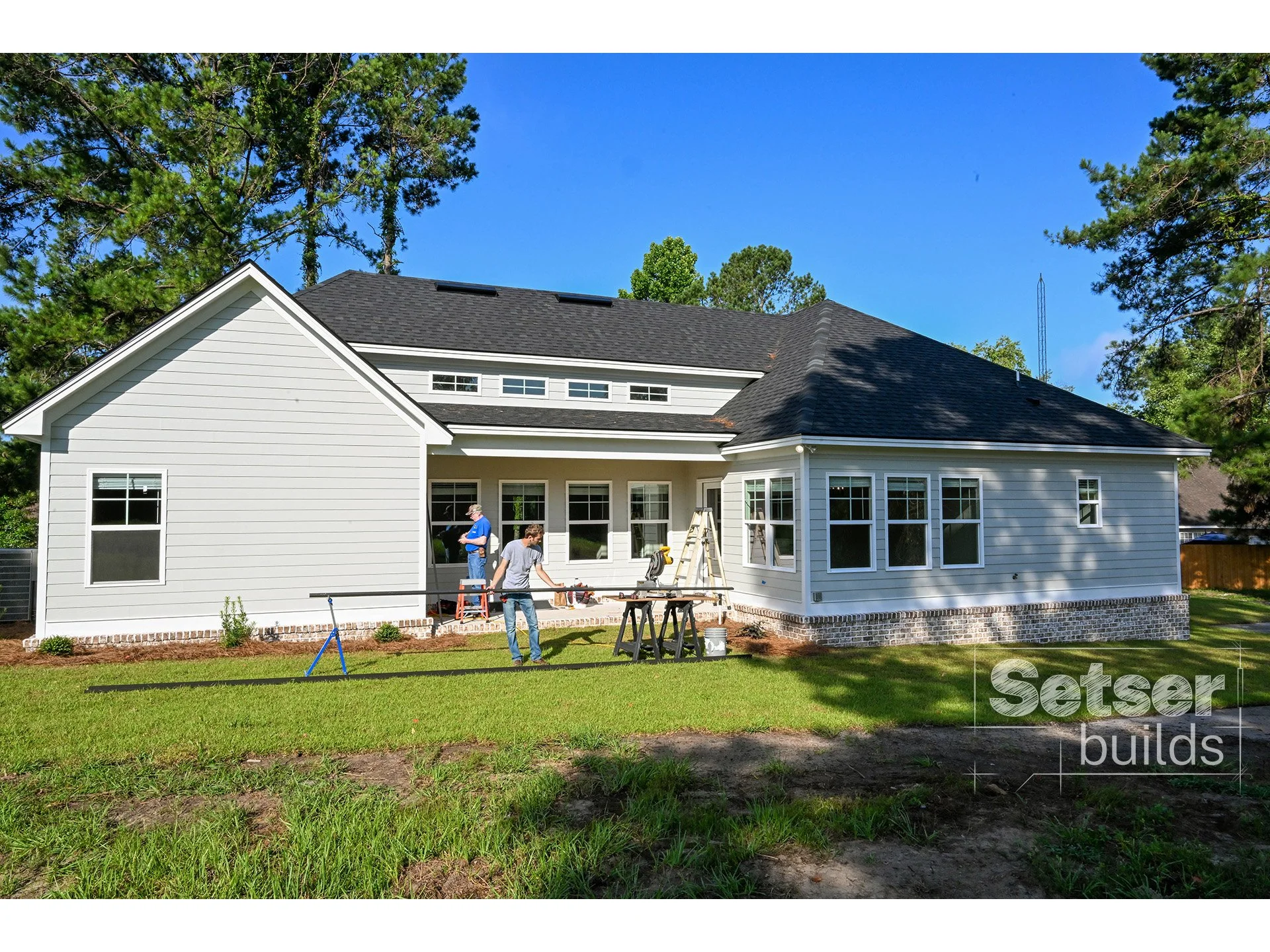 Construction workers build the back porch of a white house with multiple windows, surrounded by a green lawn and tall trees under a blue sky.