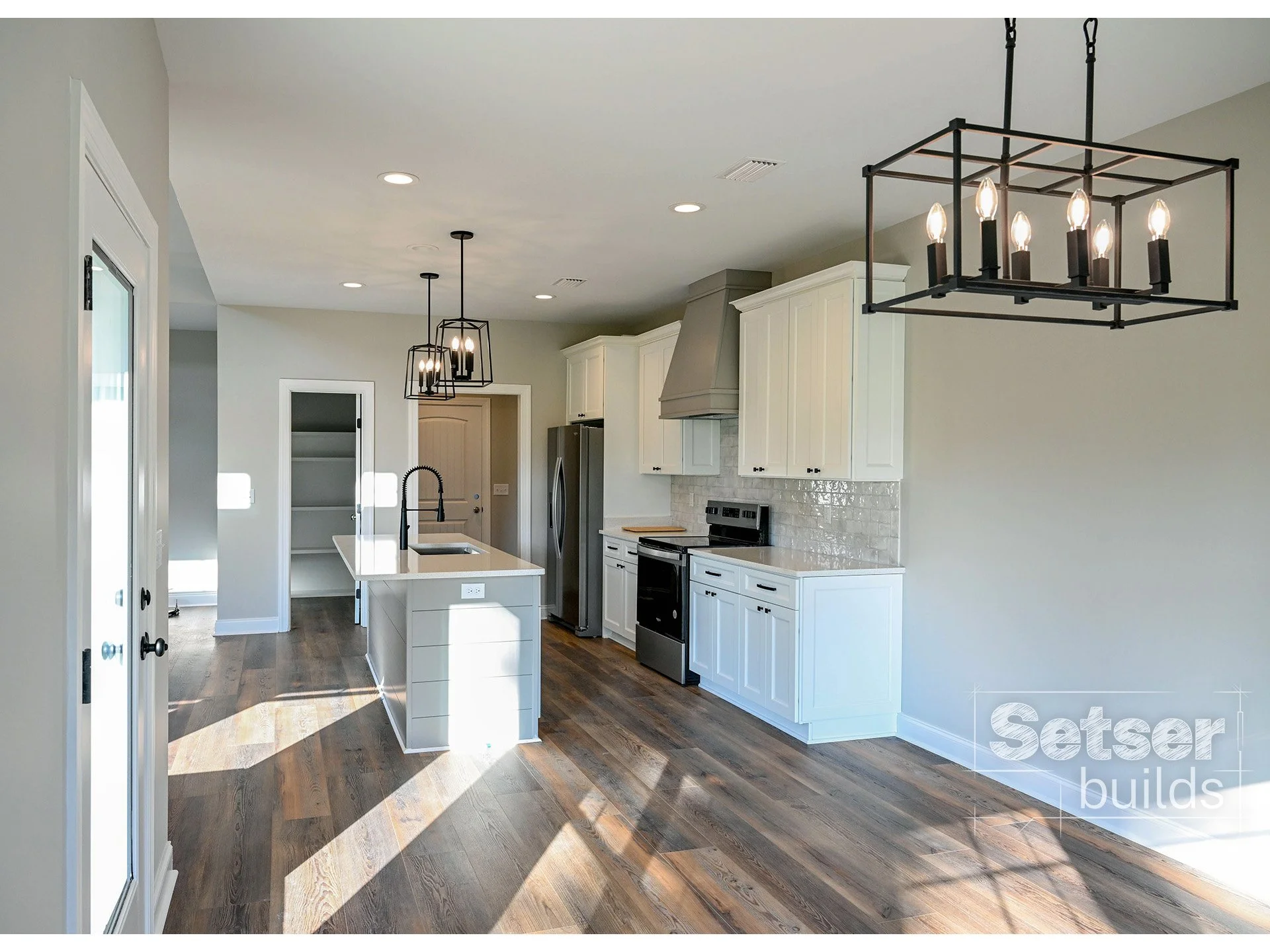 Modern kitchen with white cabinets, stainless steel appliances, and a wooden floor. There are black metal light fixtures hanging from the ceiling and sunlight coming through a door with glass panels.