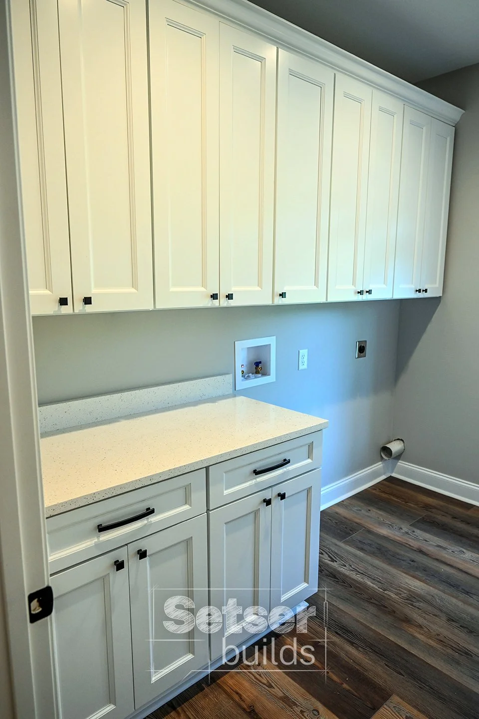 Interior view of a laundry room or utility room with white cabinets, a countertop, and an area for a washing machine and dryer, with a window and wood flooring.