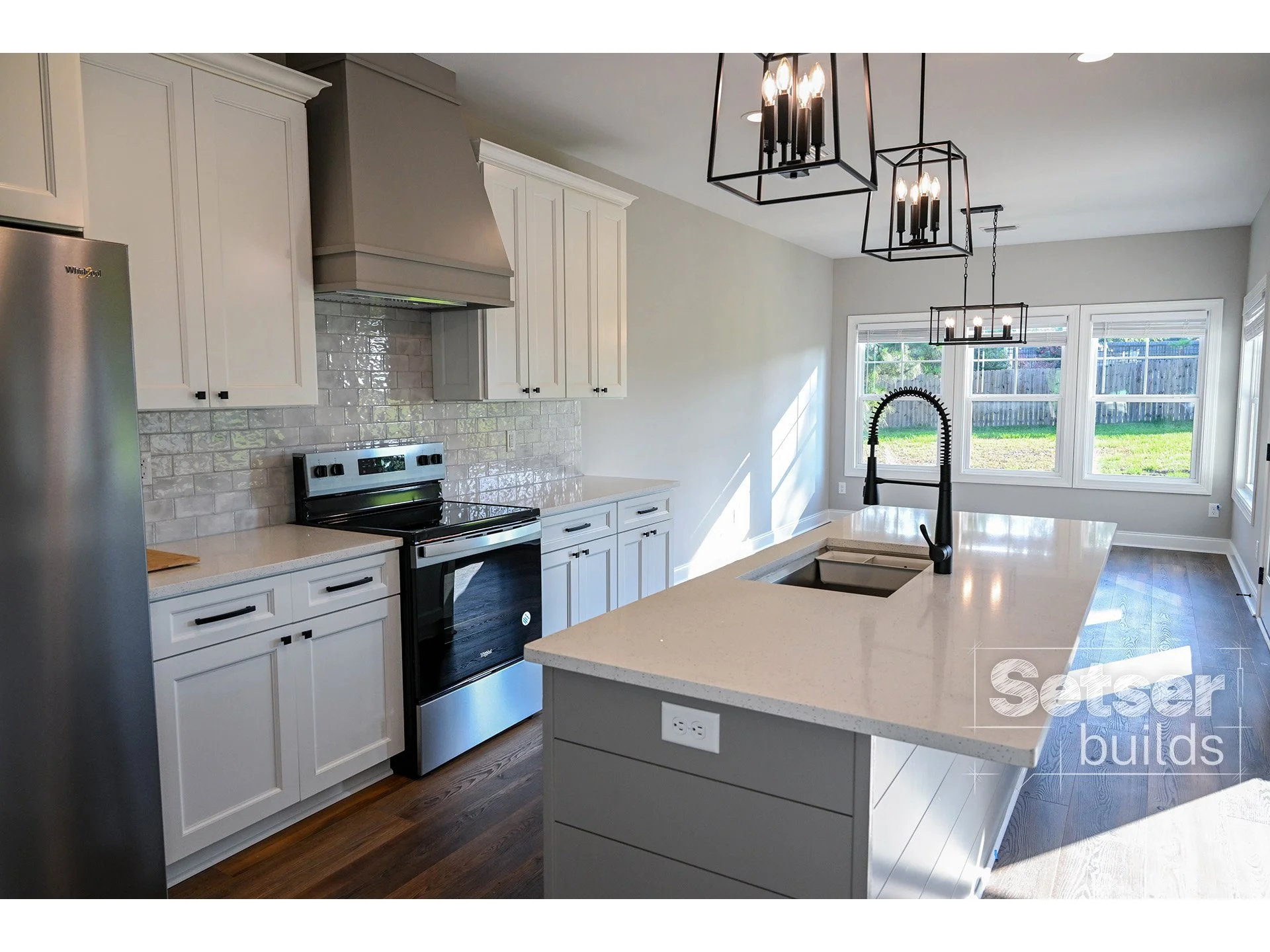 Modern kitchen with white cabinetry, black appliances, a large island with a sink and faucet, black pendant lighting, and three large windows overlooking a fenced backyard.