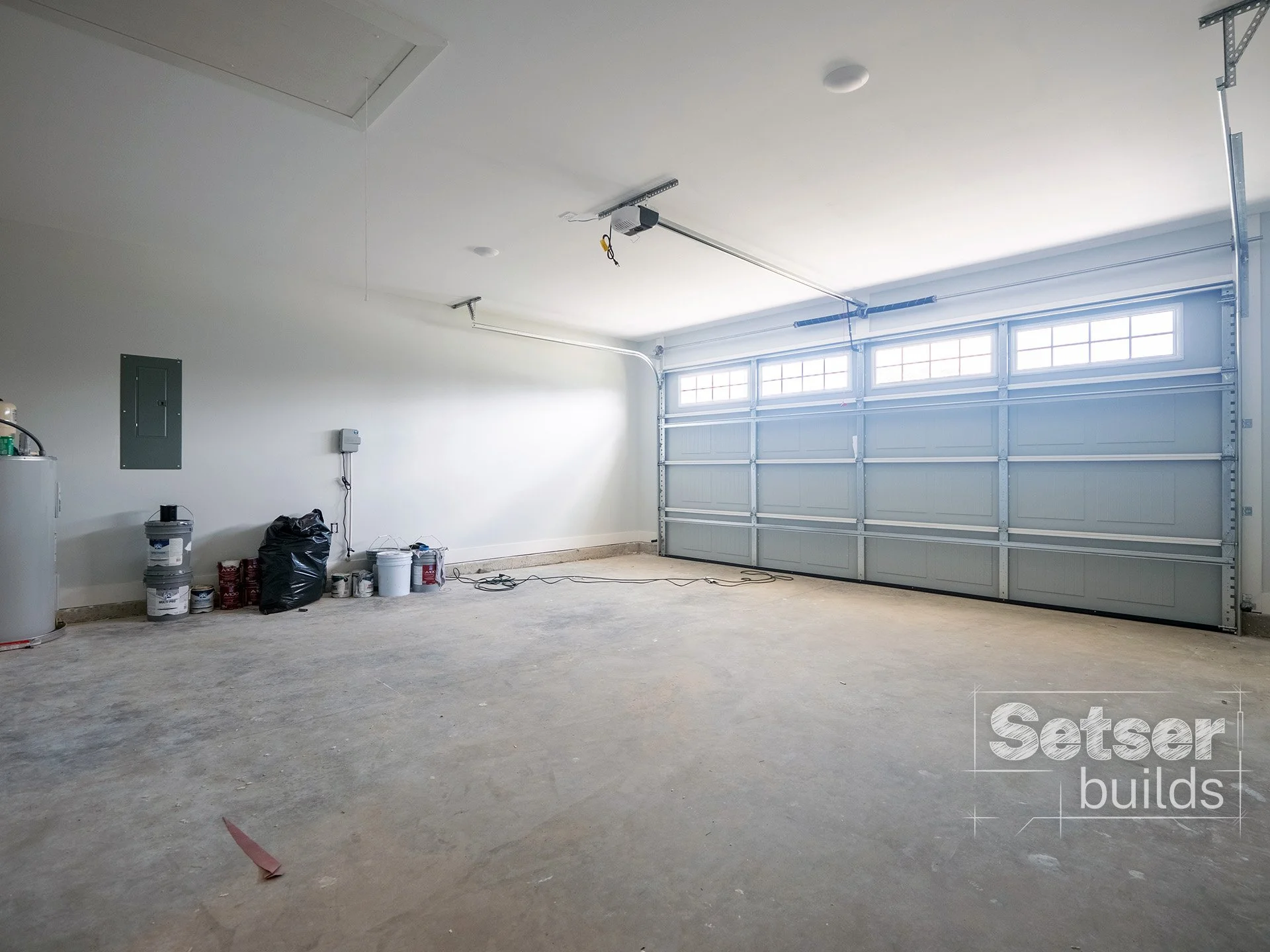 Empty garage with a closed roll-up door, some paint cans and supplies on the floor, and a utility box on the wall.