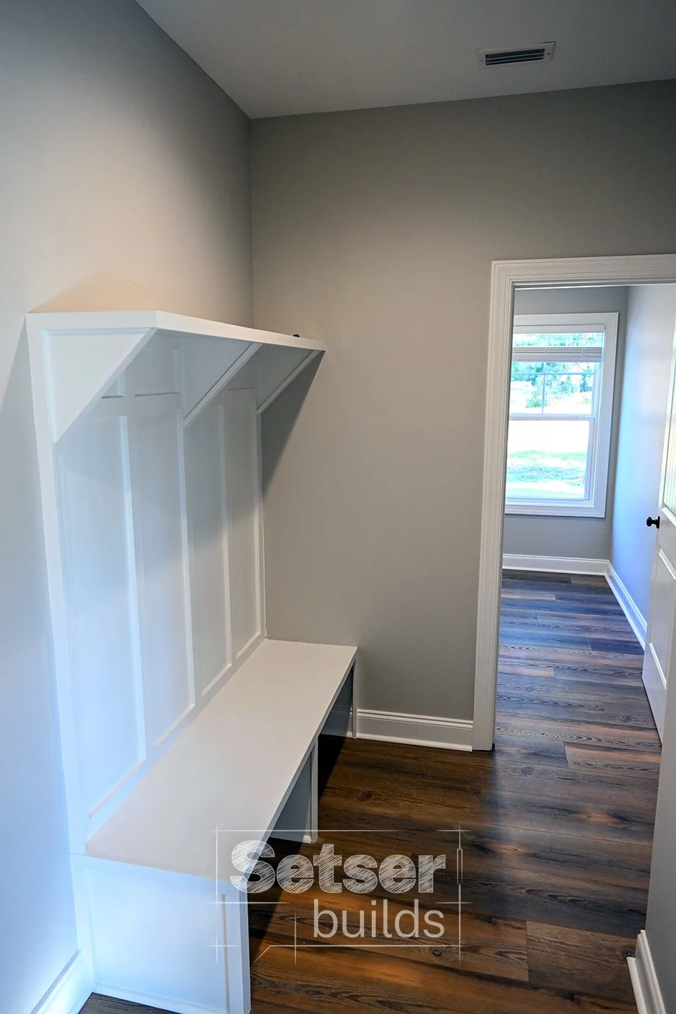 Empty laundry or mudroom with white built-in shelving and bench, gray walls, and wood flooring, leading to a bright room with a window.