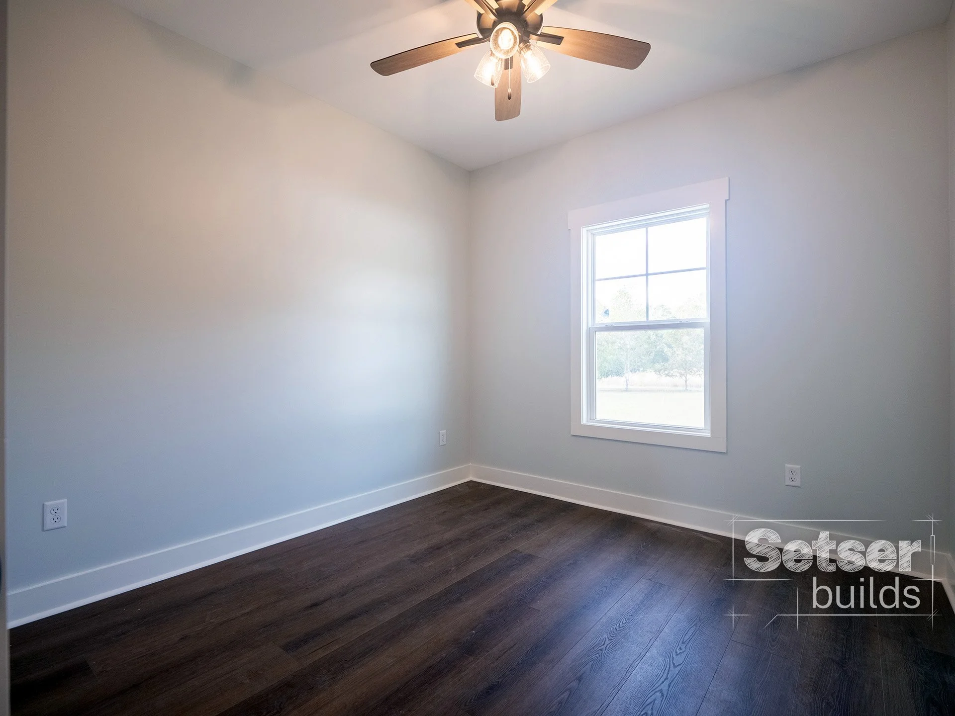 Empty room with white walls, dark hardwood floor, a window, and a ceiling fan with lights.