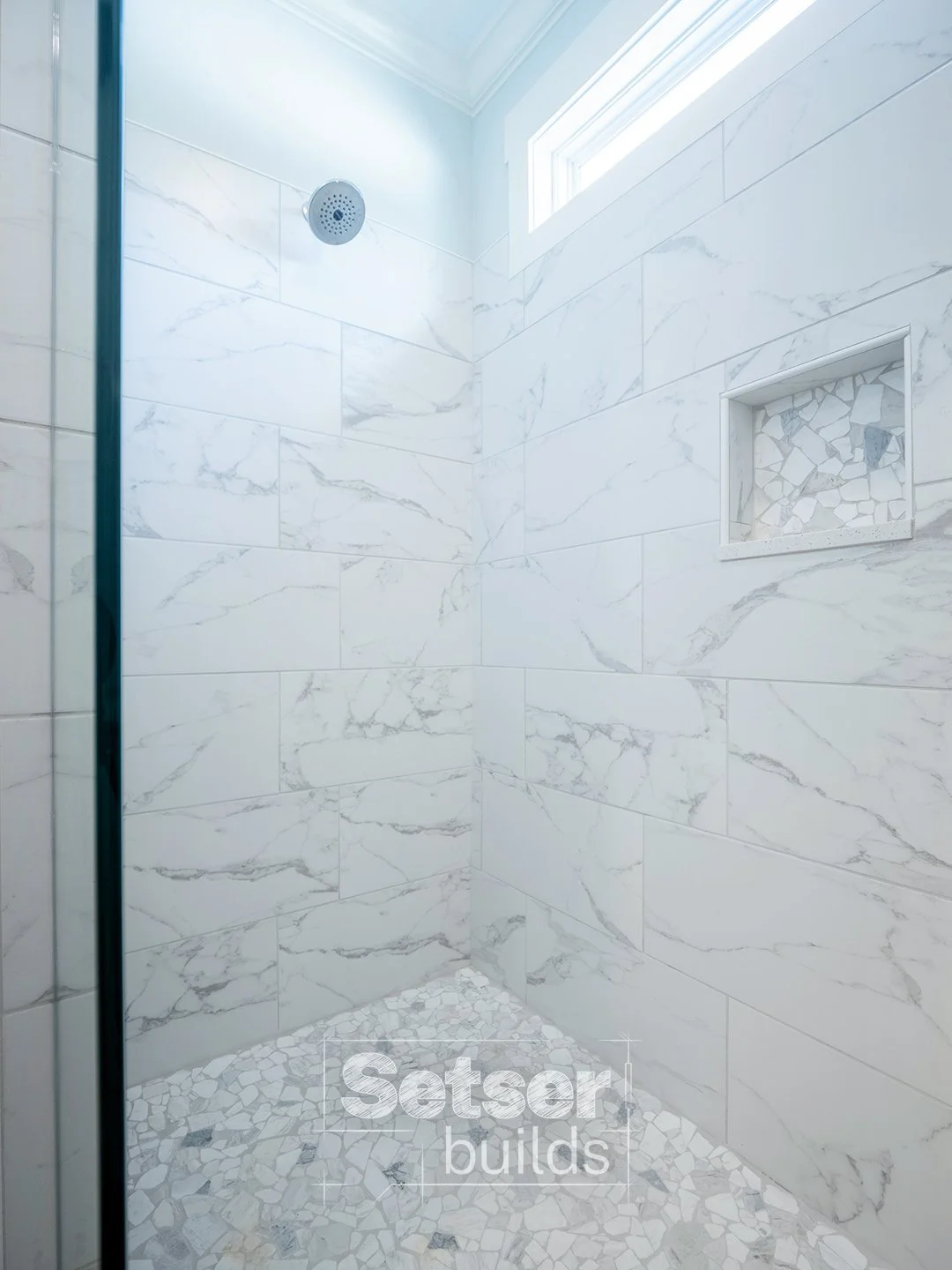 A modern shower with white marble wall tiles, a built-in niche with mosaic tiles, a rain shower head, and natural light from a high window.