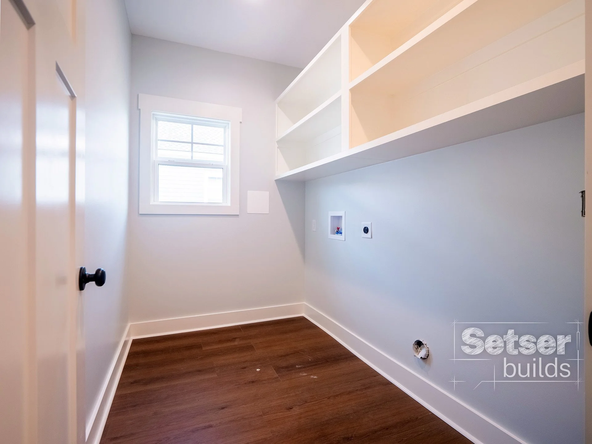 Empty laundry or utility room with white walls, a small window, and built-in shelves on the right wall. The floor is hardwood, and there are some electrical outlets and plumbing fixtures for washer and dryer. The room appears fresh and newly renovate