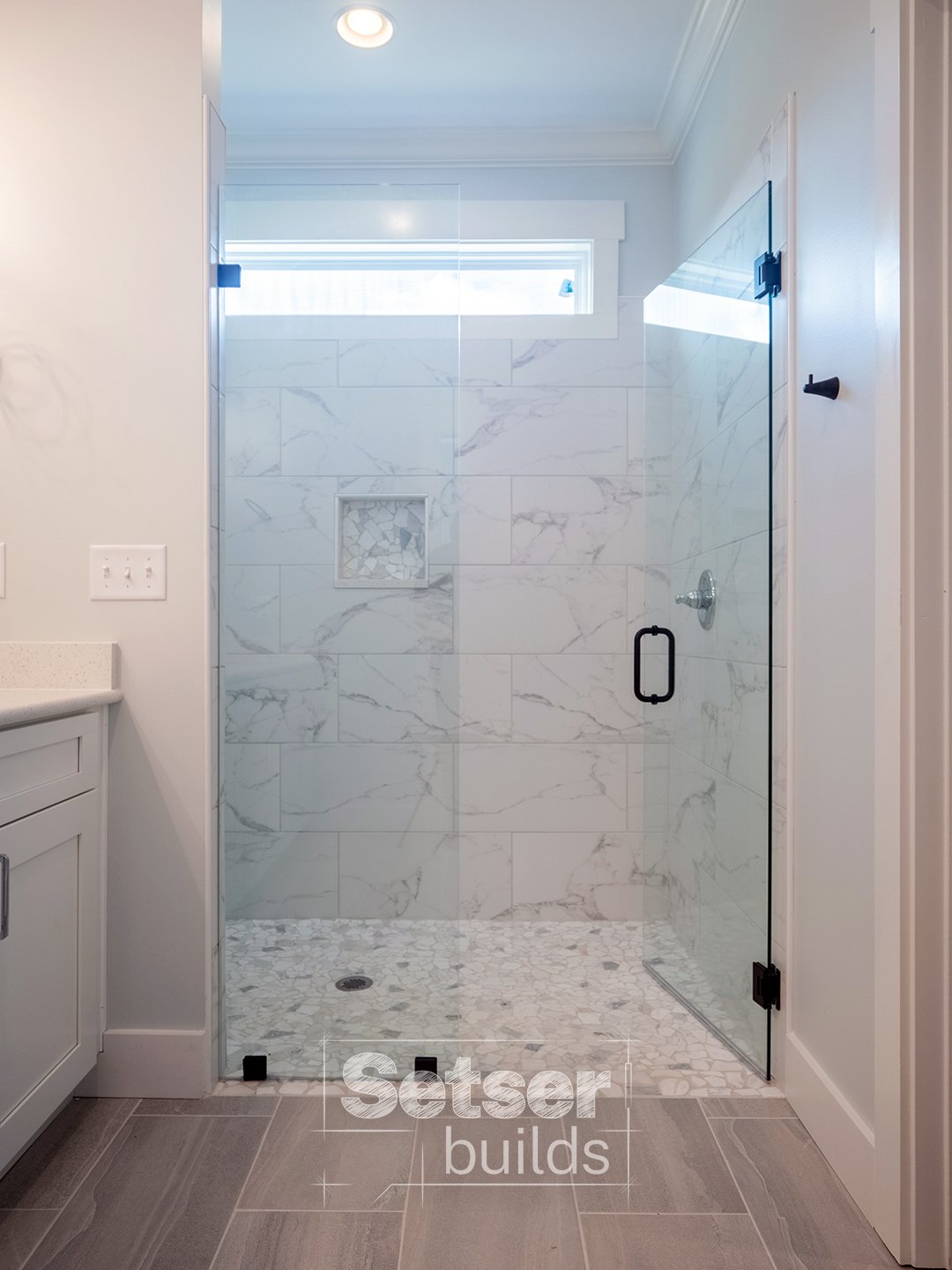 A modern bathroom shower with white marble tiles, a small window above, a glass door with a black handle, and a mosaic tile accent wall inside.