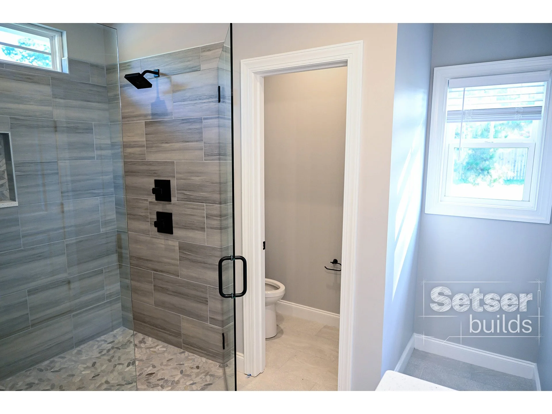 Bathroom with a glass-enclosed shower area with tiled walls and pebble flooring, a small window, and a separate toilet room with a door.