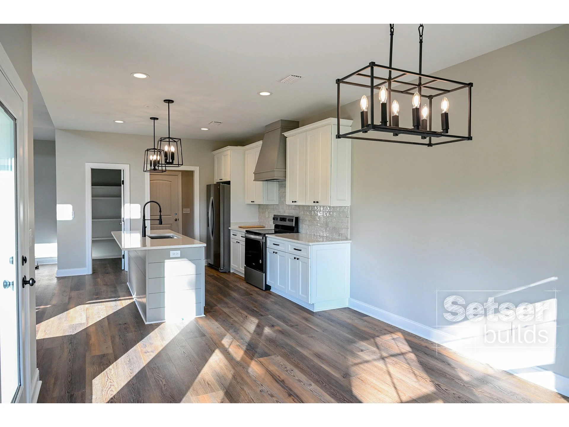 Modern kitchen with white cabinets, stainless steel appliances, hardwood flooring, black metal light fixtures, and a large empty wall.