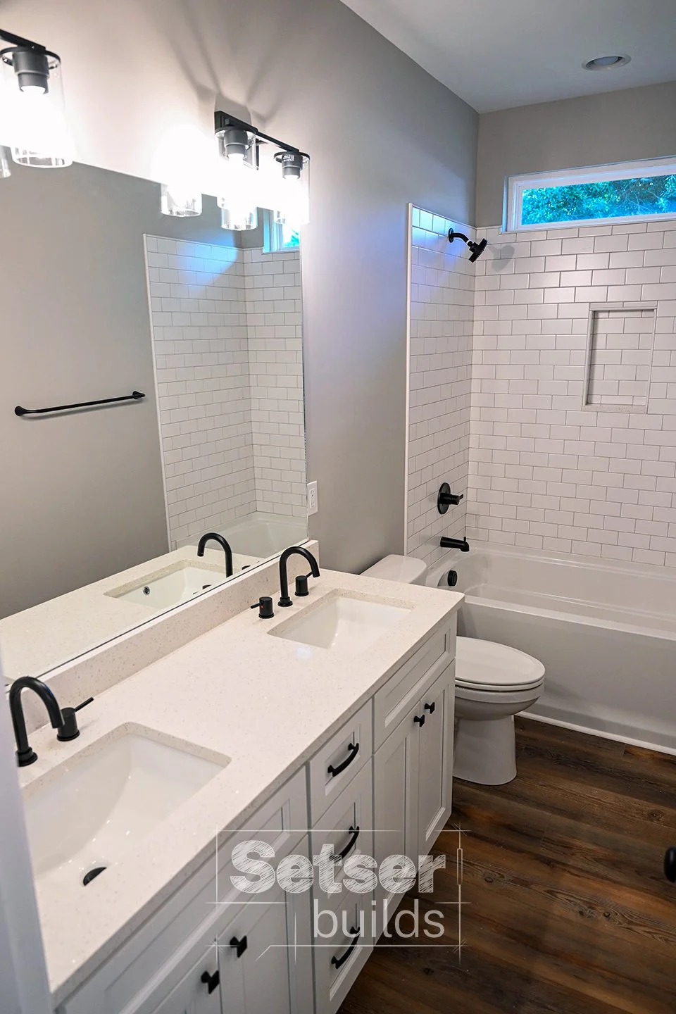 Bathroom with white double sink vanity, black fixtures, large mirror, and a shower with white subway tile, black fixtures, a small window, and a bathtub.