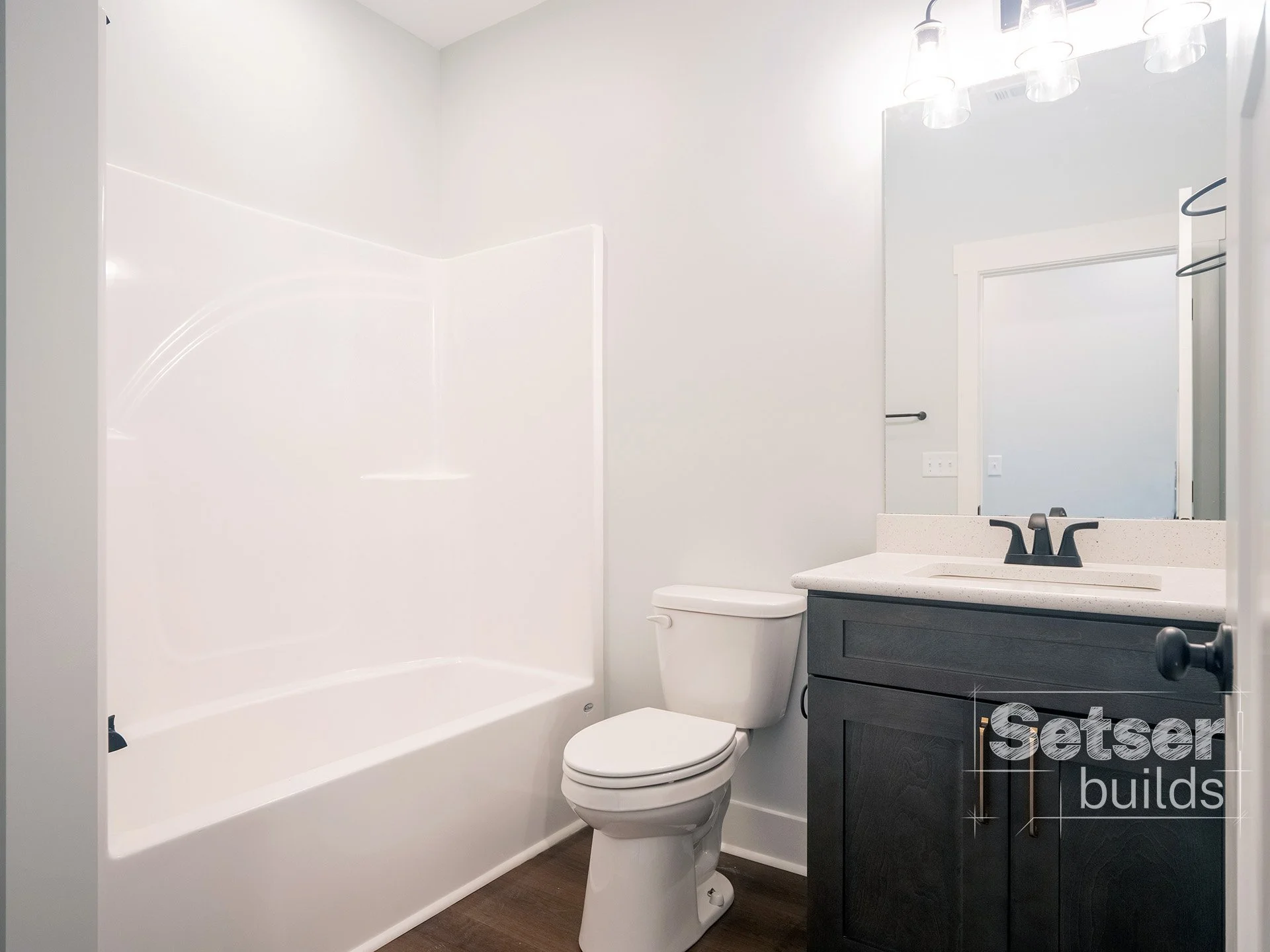 A modern bathroom with a white bathtub, toilet, black vanity with a white countertop, mirror, and three light fixtures above the mirror.