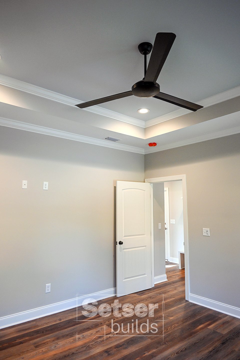 Empty room with beige walls, white trim, dark wood flooring, a ceiling fan, and an open door leading to another space.