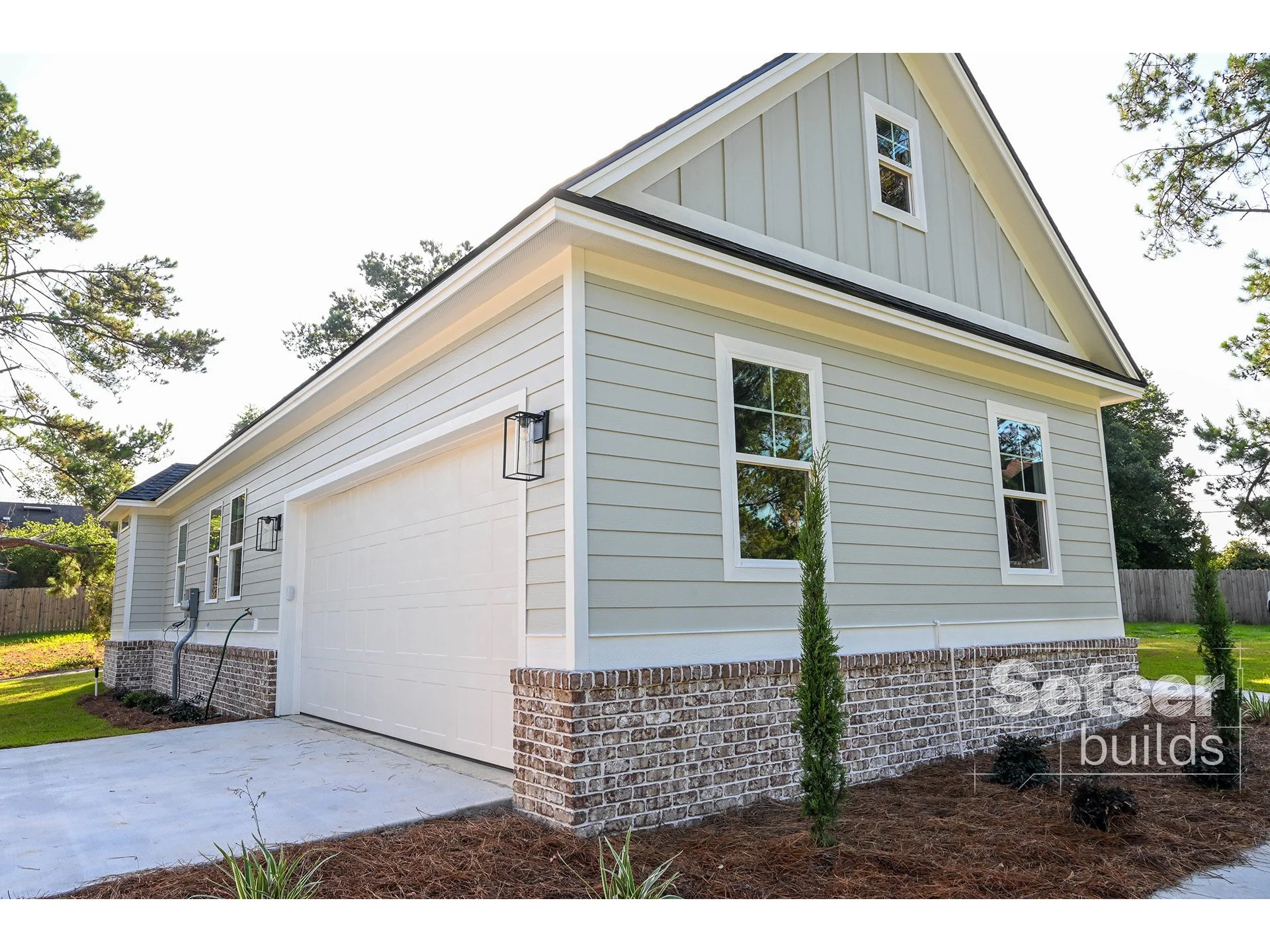 Modern two-story house with white siding and brick foundation, featuring a garage door, multiple windows, and landscaped yard with small trees.