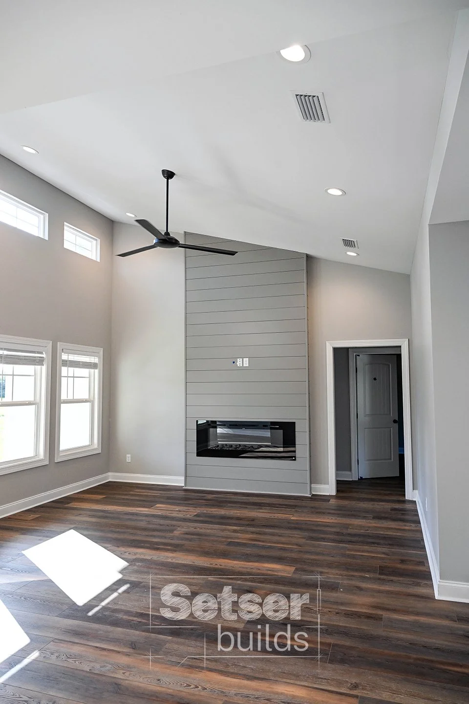 Empty living room with gray walls, wood flooring, a modern black ceiling fan, multiple small windows near the ceiling, a built-in fireplace, and a door leading to another room.