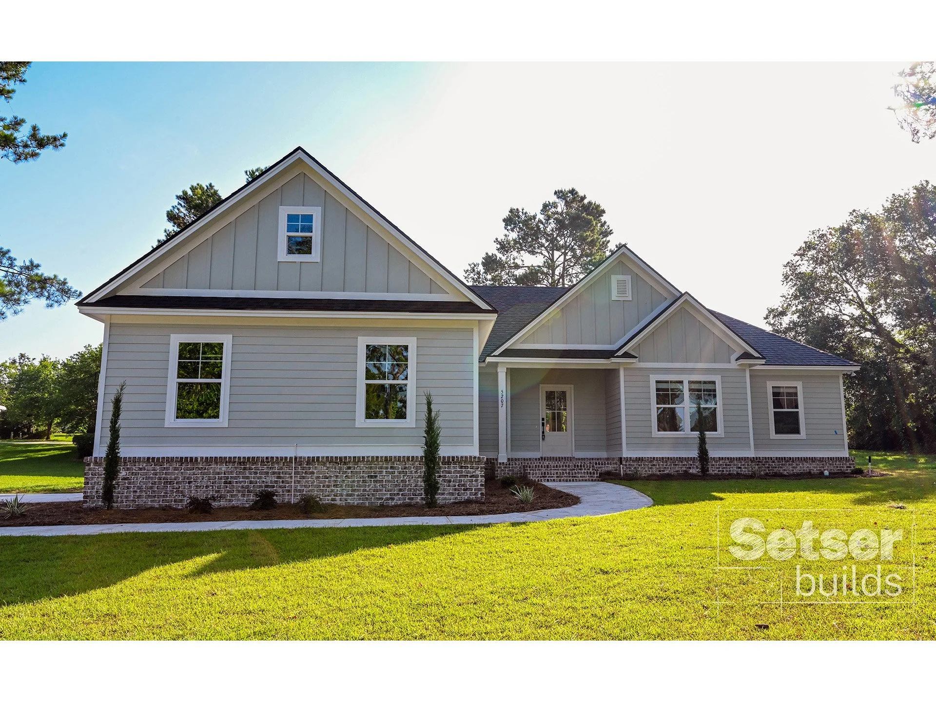 Modern two-story house with white siding, brick foundation, multiple gables, and large windows, surrounded by a green lawn and trees.