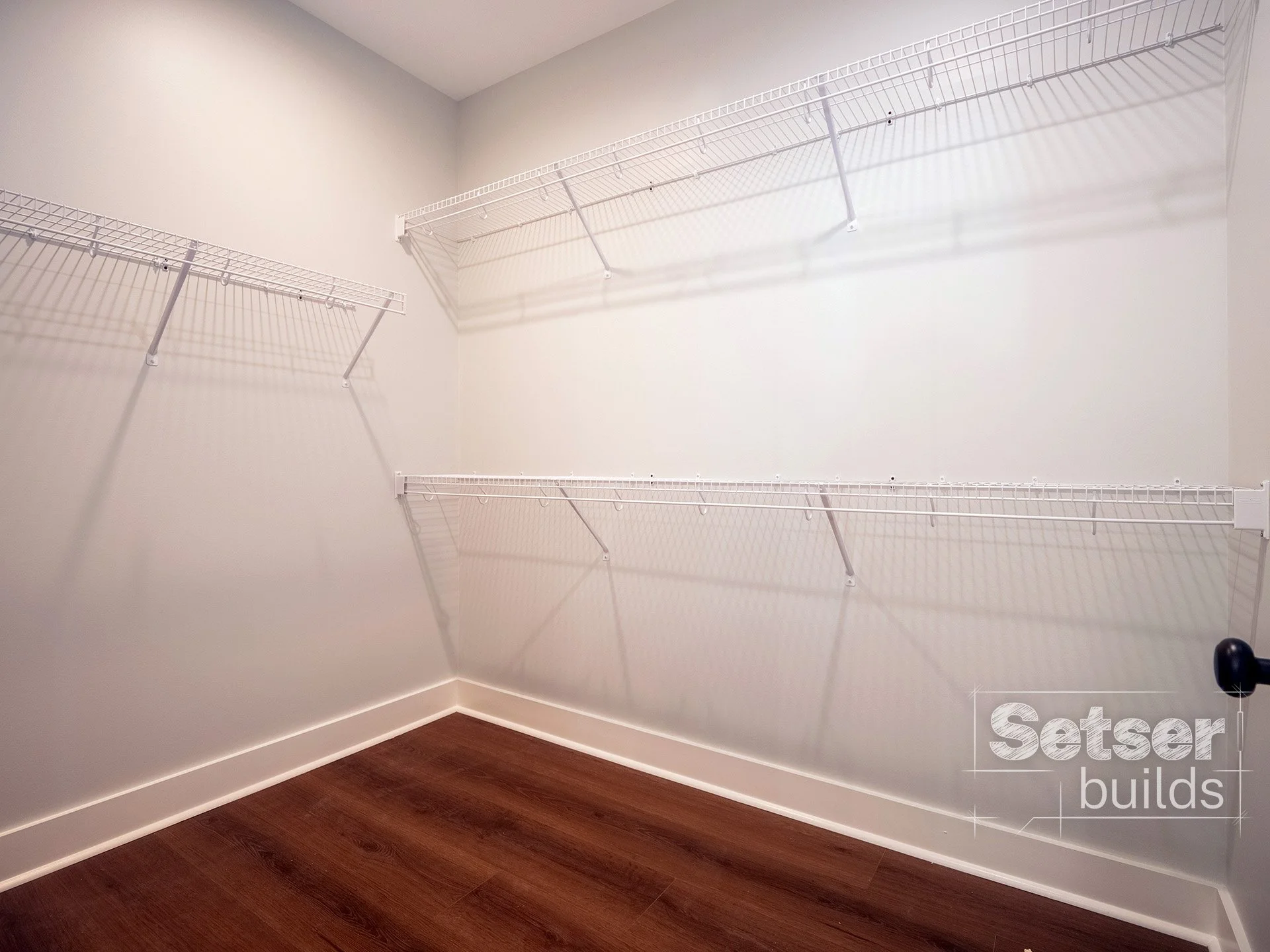 Empty walk-in closet with white wire shelves on white walls and hardwood flooring.