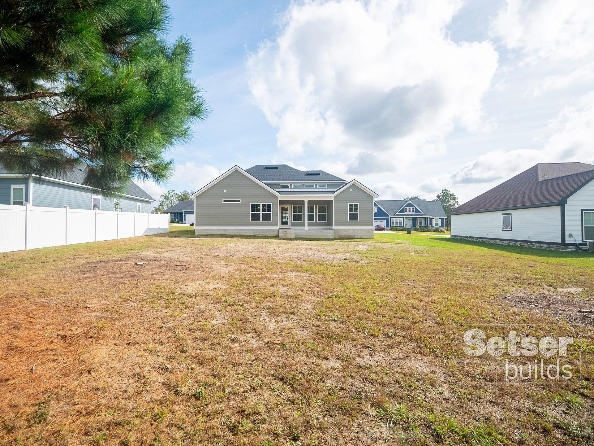 The image shows a backyard with a new house at the center, surrounded by other houses on both sides. The yard has patchy grass and dirt, and a large tree on the left side with green pine branches. The sky is partly cloudy with some sunshine.