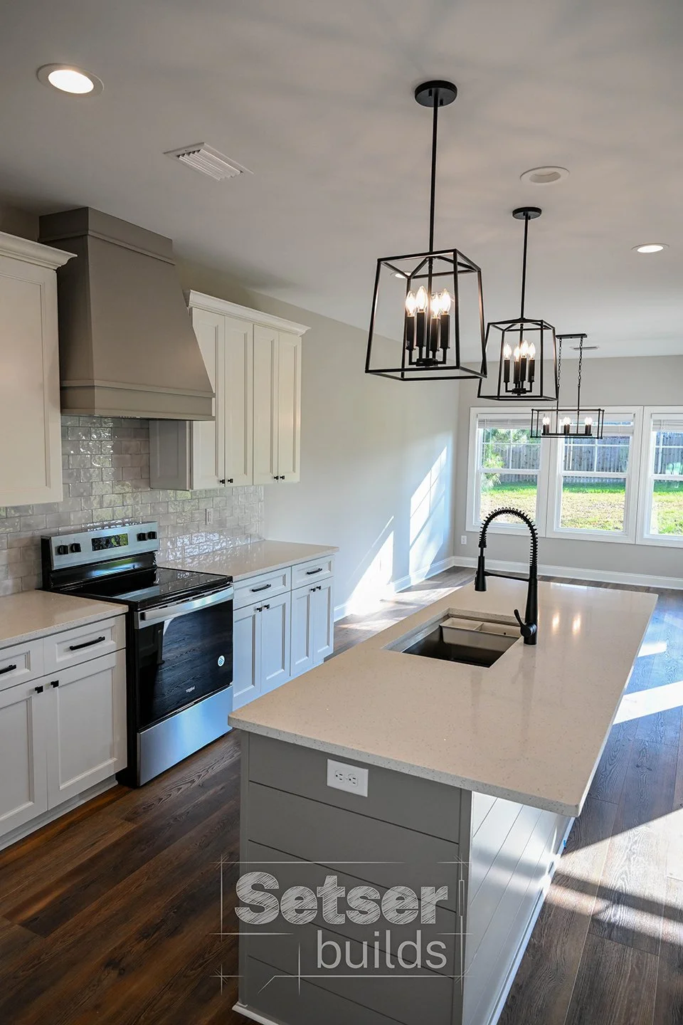 Modern kitchen with white cabinets, a black stove, a large island with a beige countertop, a black faucet, pendant lighting, and large windows letting in natural light.