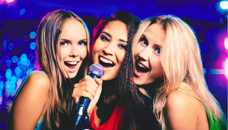 Three women singing and having fun at a karaoke night with colorful lights in the background.
