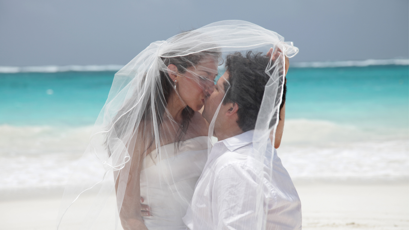 Bride and groom share a romantic kiss on the beach, with the bride’s veil draped over both their heads.
