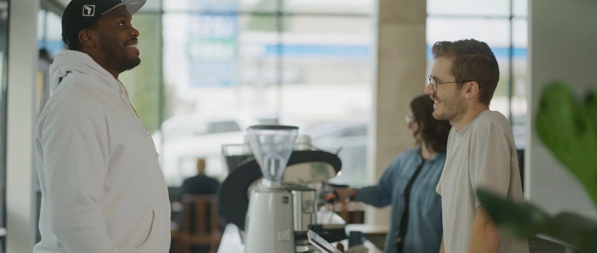 A man walking into Liminal and ordering a coffee from the barista. They are smiling and facing each other, with a coffee grinder and a woman working in the background.