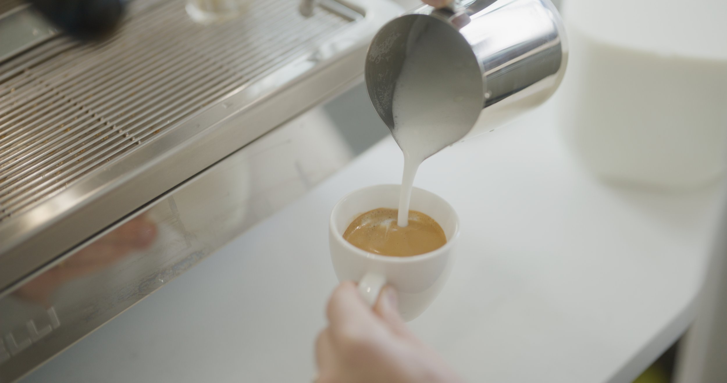 Barista pouring steamed milk into a cup of espresso coffee on a white counter.