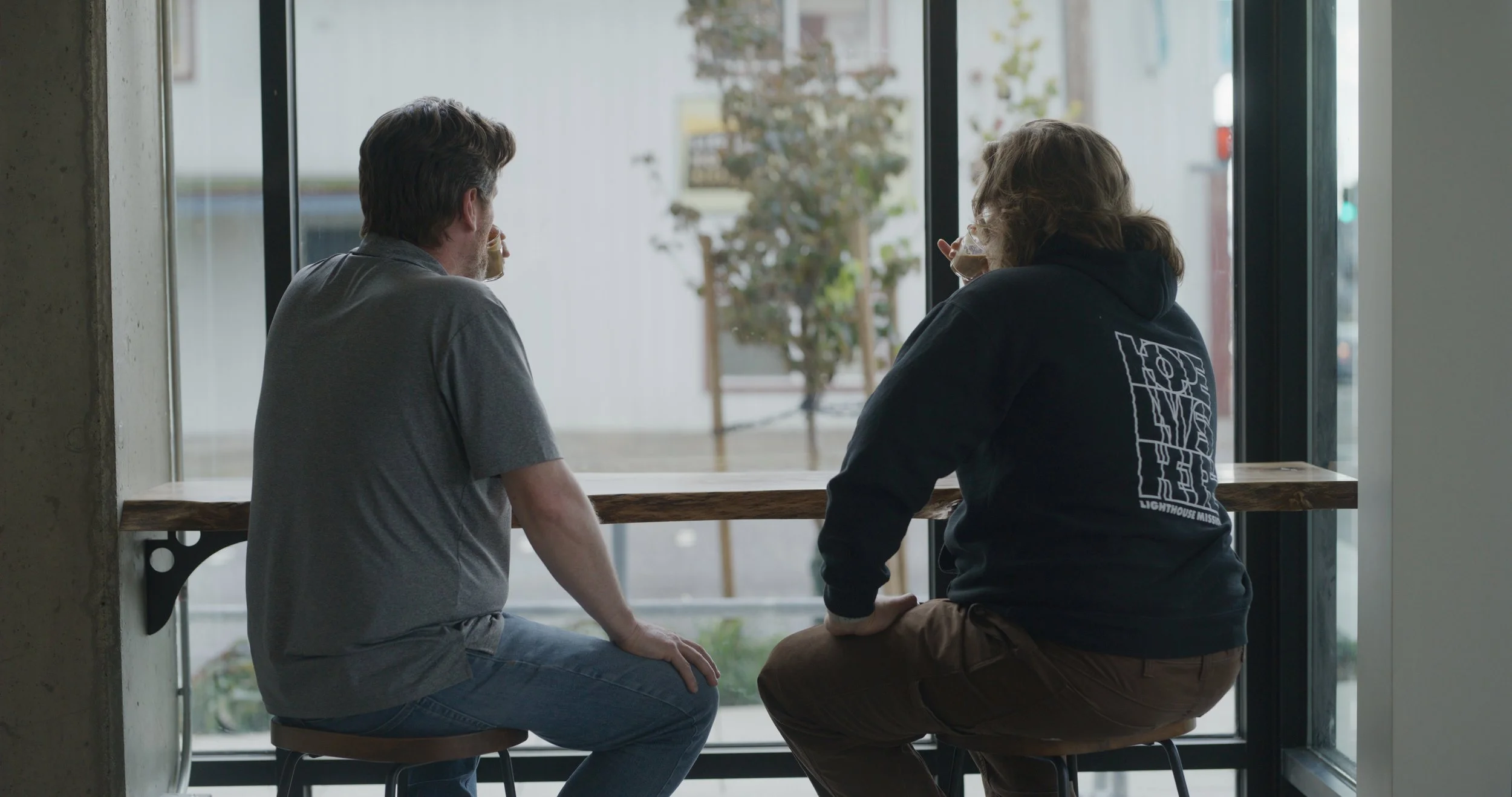 Two men sitting at a wooden counter by a large window, drinking coffee and talking.