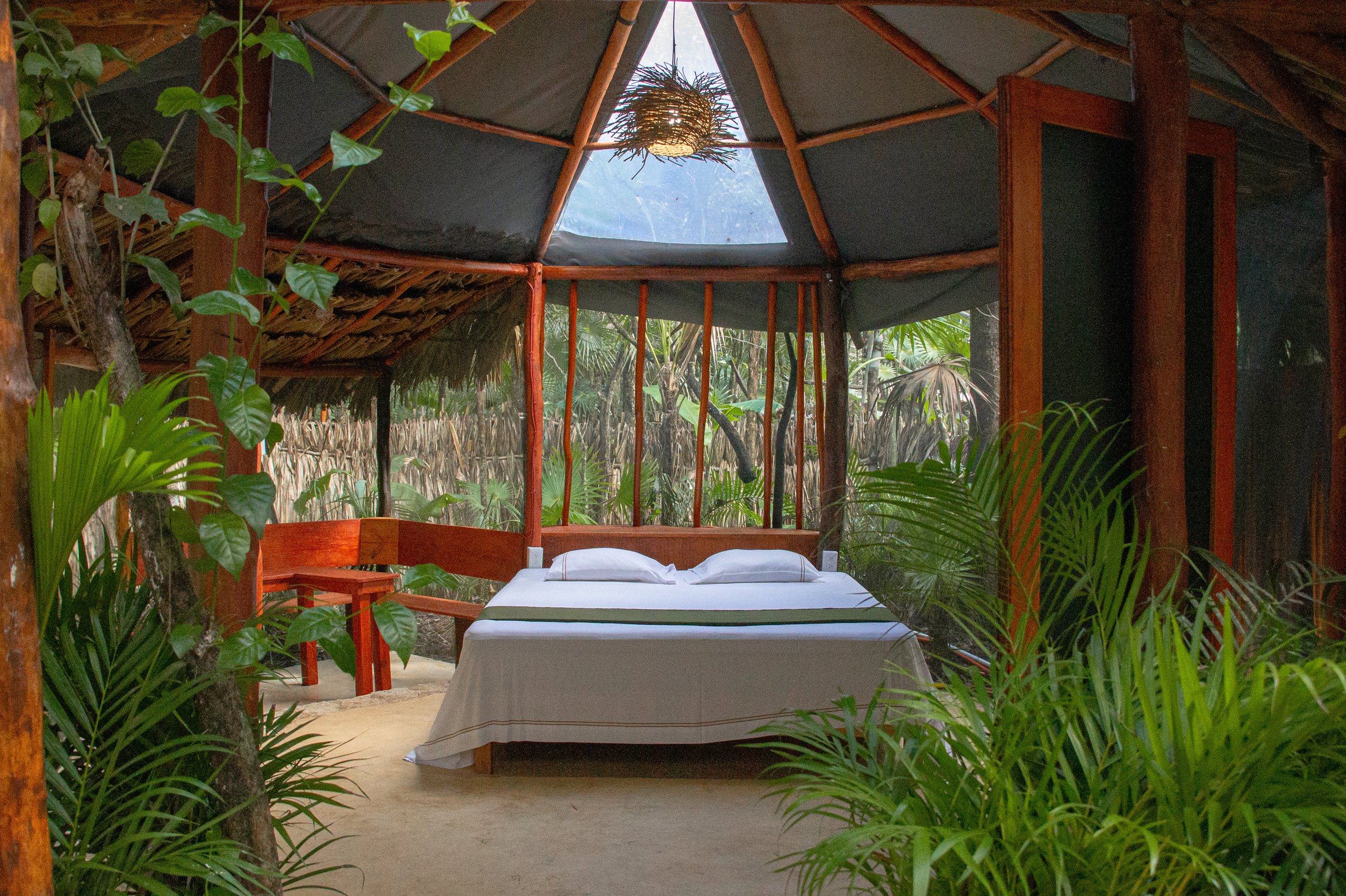 A cozy, rustic bedroom set in a jungle-style hut with a wooden bed, white linens, and green accents. Surrounded by lush greenery and with a thatched roof, the room features a skylight with a woven light fixture hanging from the ceiling.