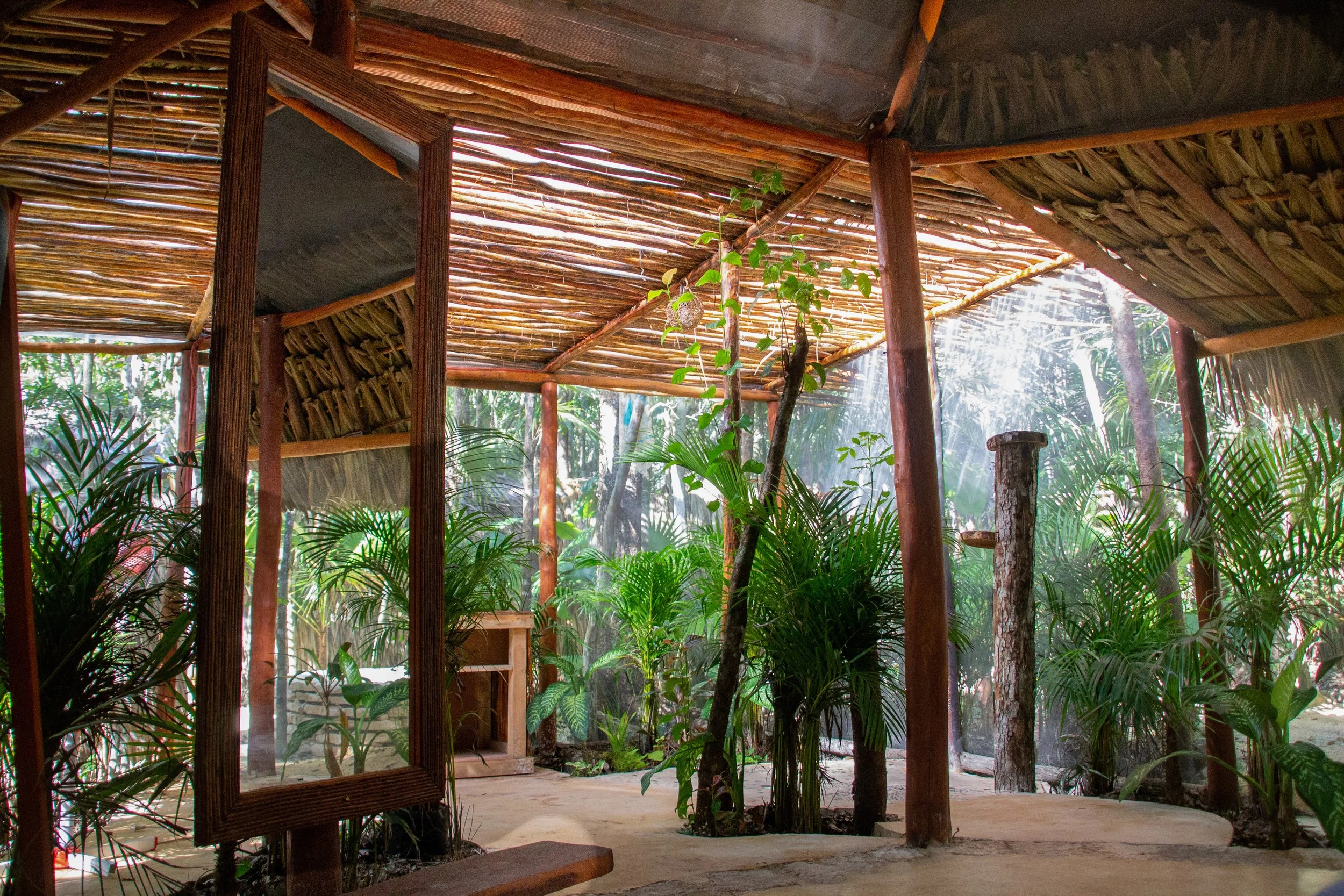 Interior of a lush, rustic tropical hut with wooden poles and a thatched roof, surrounded by dense green plants and trees, sunlight filtering through the foliage.