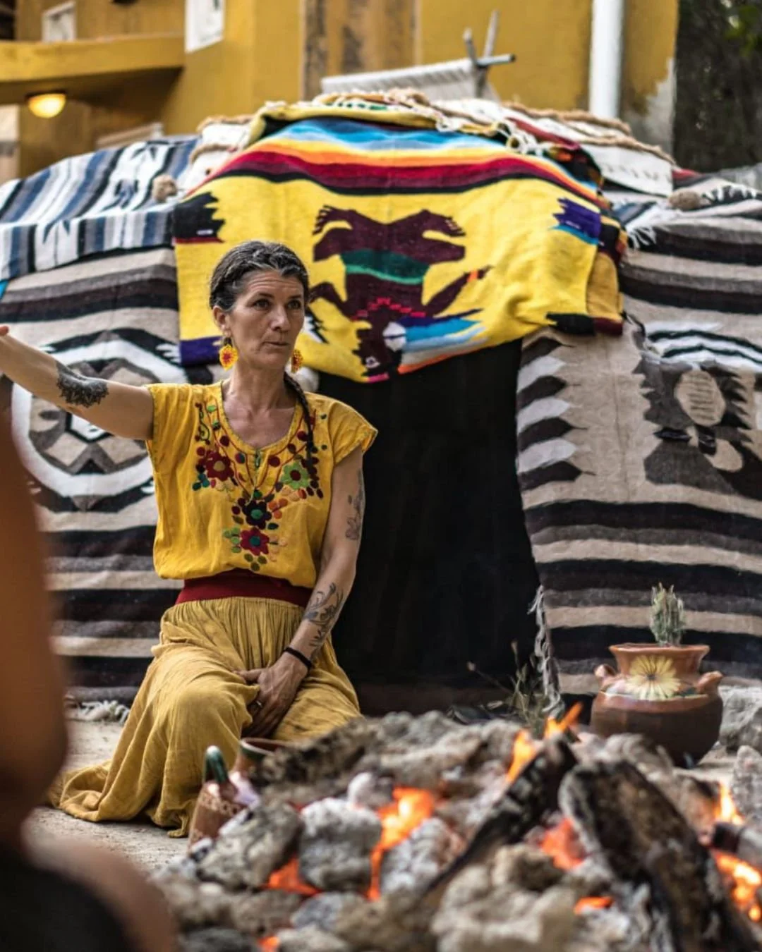 A woman dressed in traditional Mexican clothing sits by a small fire with a decorative pottery piece nearby, with colorful textiles and blankets hanging behind her.