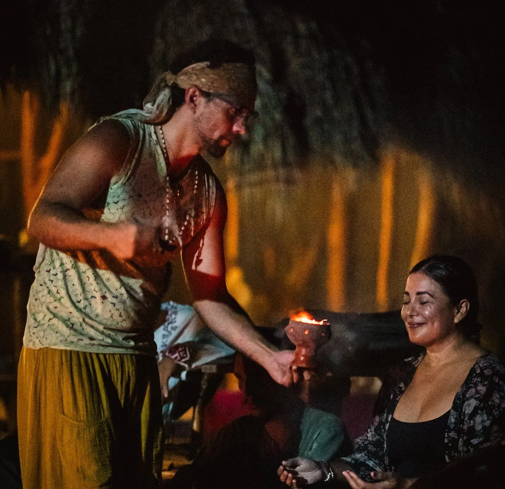 A man in bohemian style with a bandana on his head offers a woman a traditional object during a nighttime outdoor gathering. The woman, with dark hair and wearing a dark top and floral shawl, is smiling with closed eyes, sitting in front of a motorbike.