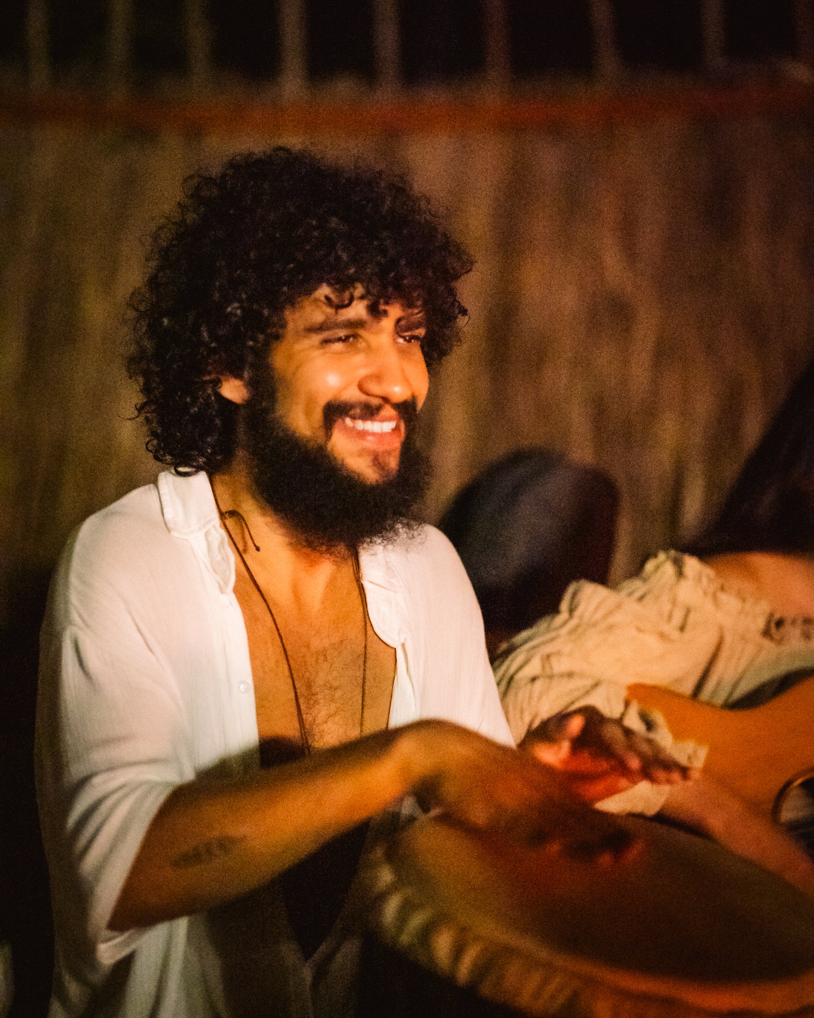 A man with curly hair and a beard smiling in a dimly lit setting, sitting at a table with a guitar nearby.