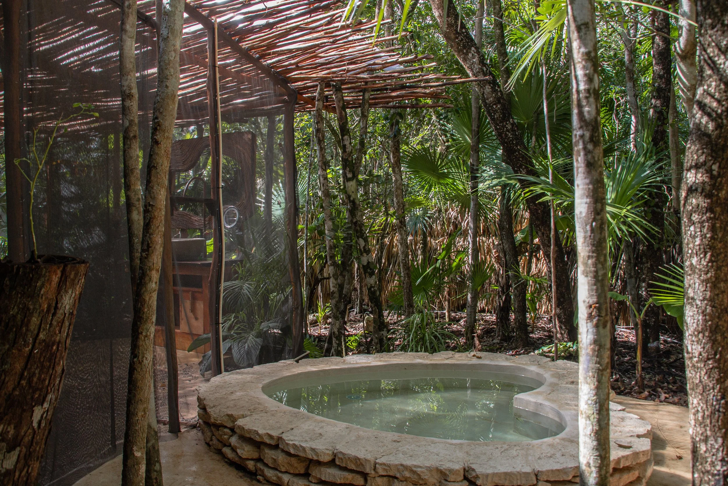 A small, circular hot tub or plunge pool made of stacked stones is situated in a lush, tropical forest with dense greenery and trees surrounding it.