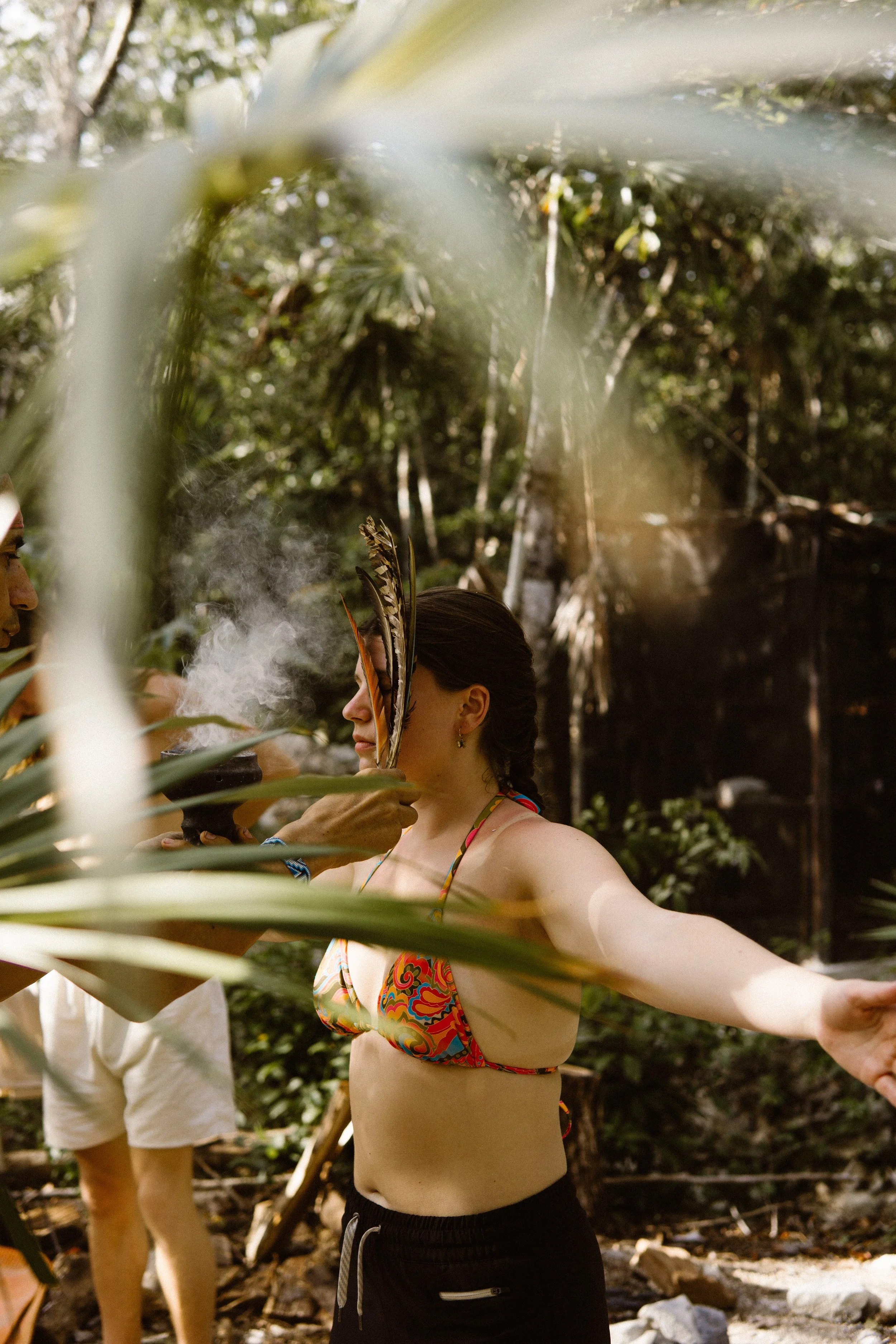 A woman in a colorful bikini top with outstretched arms standing outdoors in a lush forest during a ceremony, with a person holding a feathered staff or object near her face.