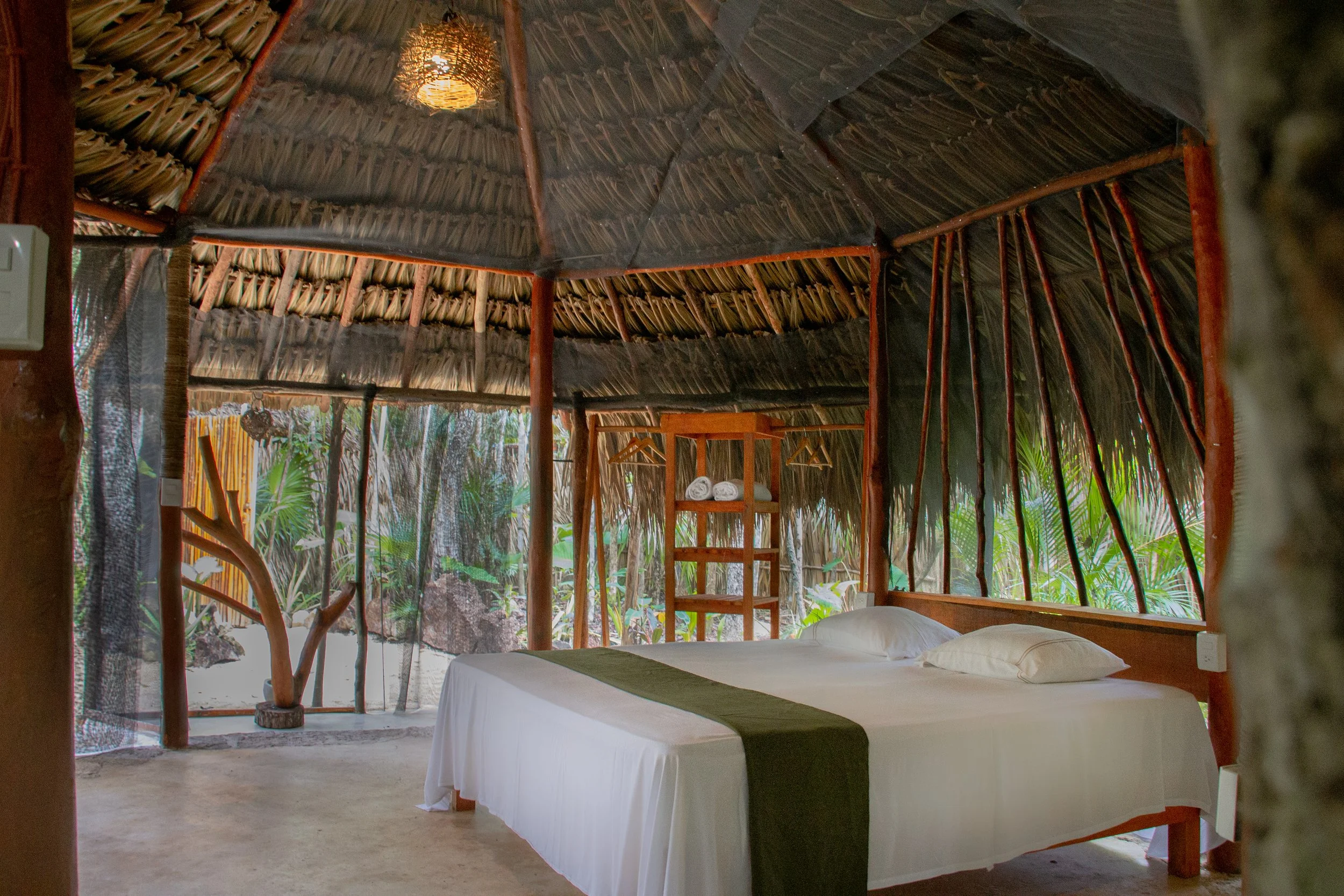 A rustic open-air bedroom with a bamboo and thatch roof, a white bed with a green runner, overlooking a lush tropical garden.