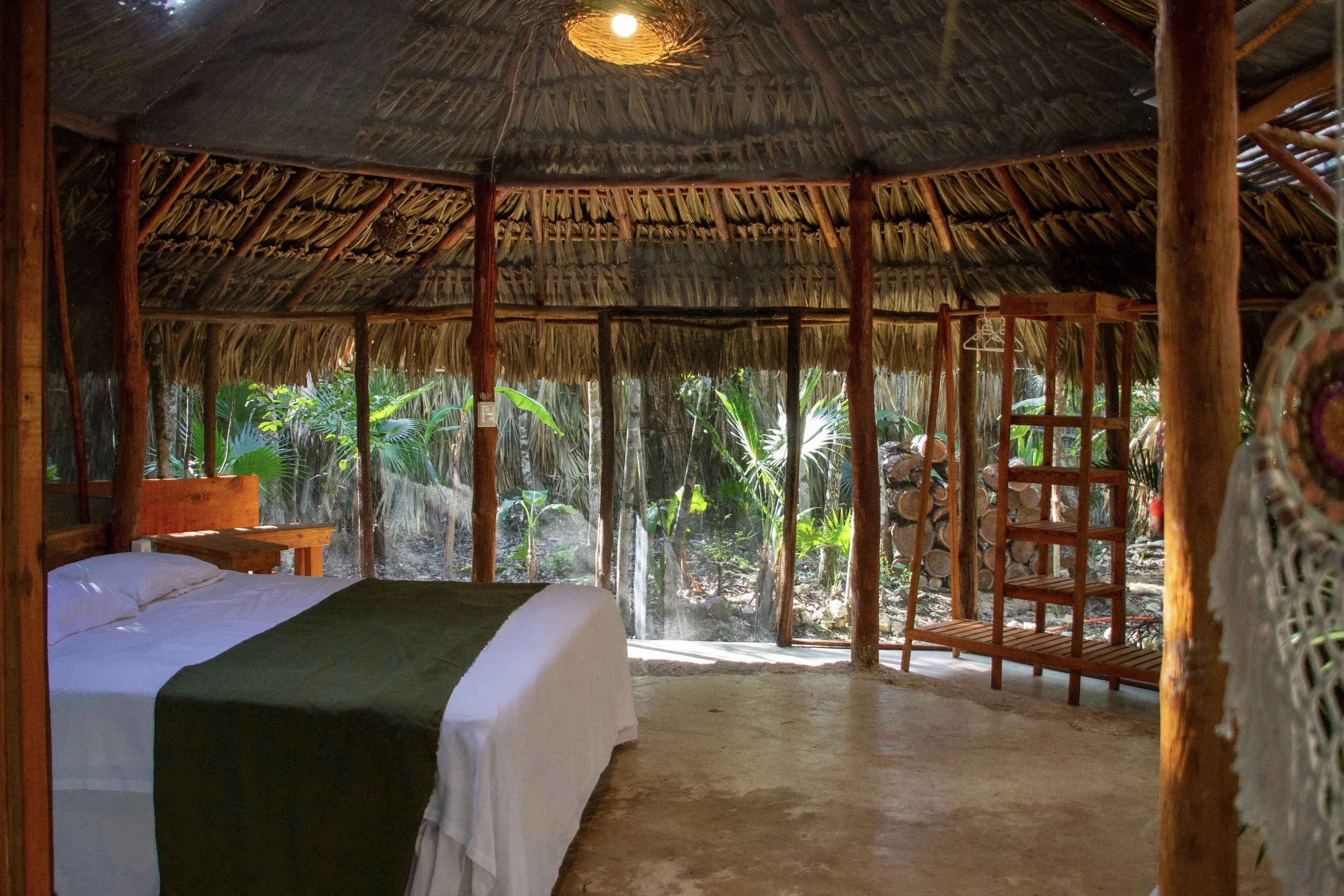 Interior of a rustic, open-air hut with a bed, surrounded by lush tropical vegetation.