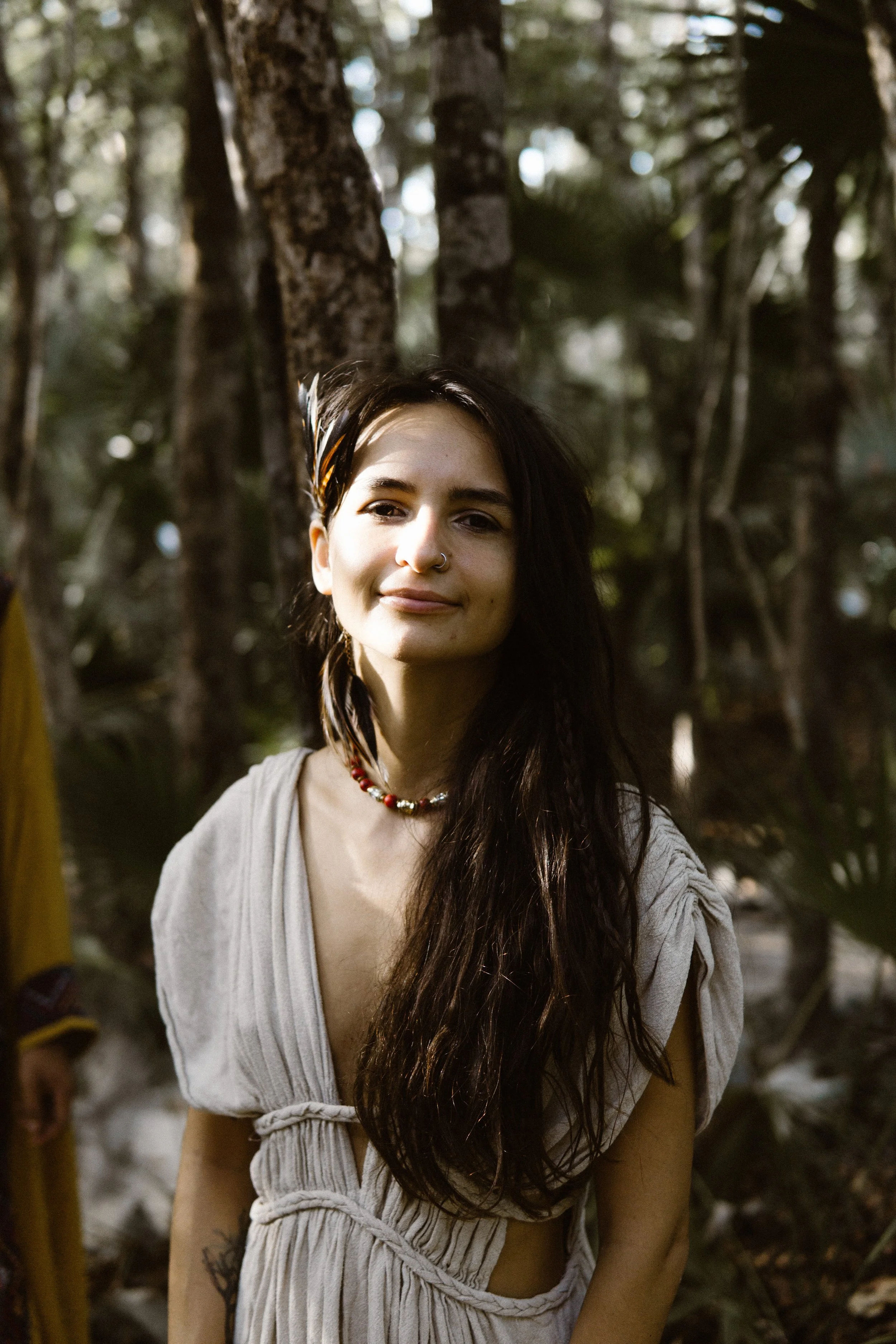 A young woman with long dark hair, wearing a beige dress and a beaded necklace, stands outdoors in a wooded area, smiling softly.