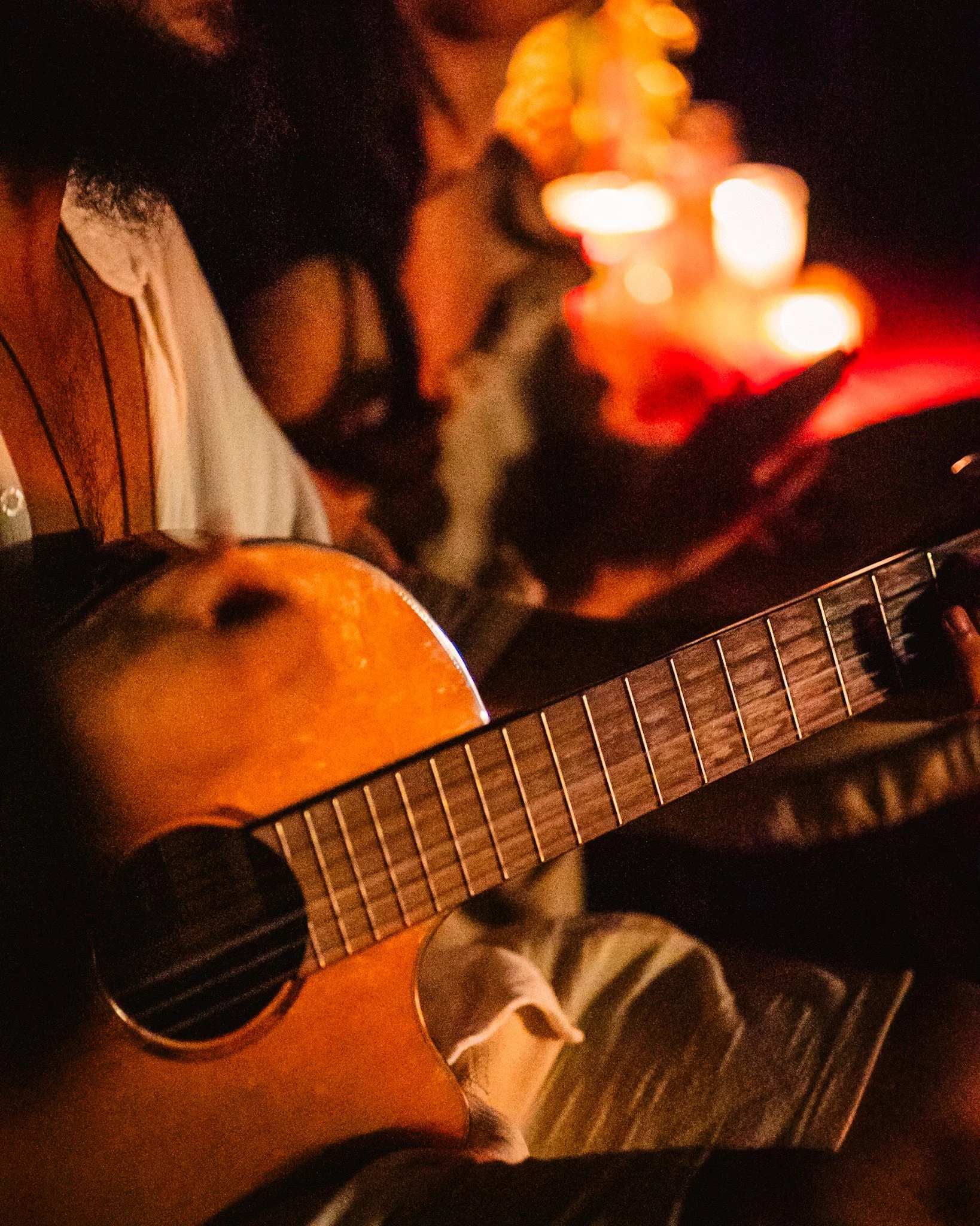 Person playing an acoustic guitar, with candles and a person using a phone in the background, in a dimly lit setting.