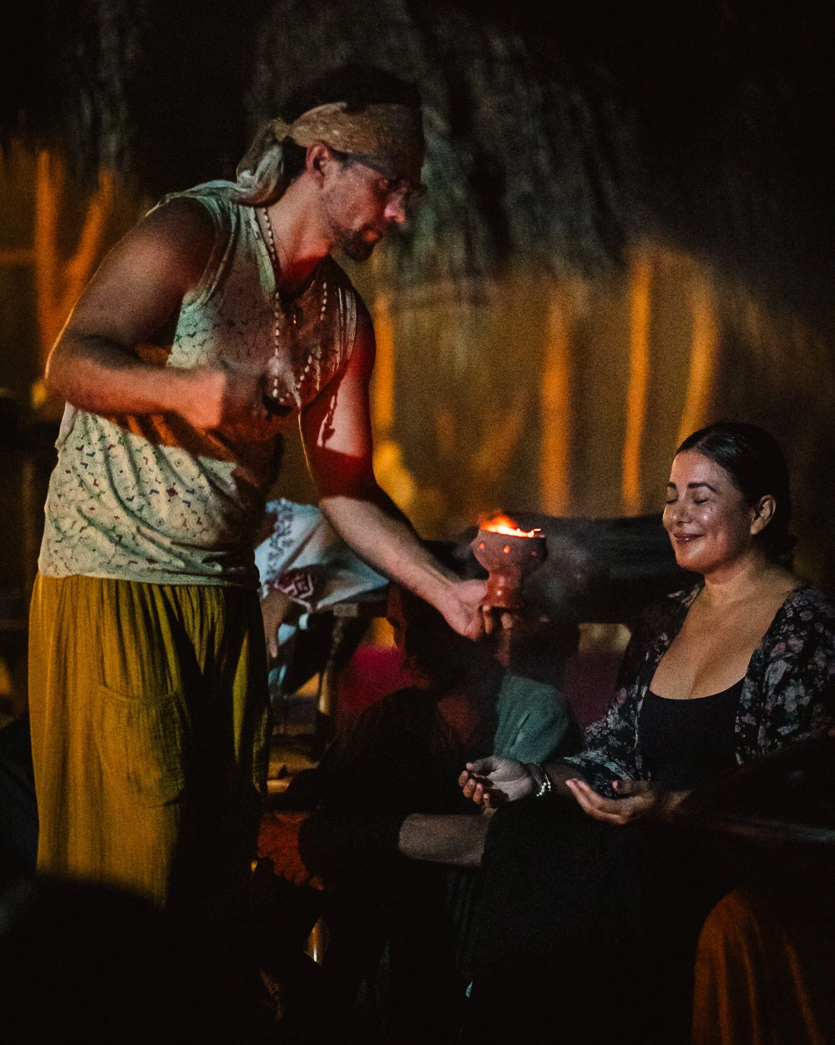 A man holding a lit torch presenting it to a seated woman with her eyes closed, in a dark environment with a cave-like background.