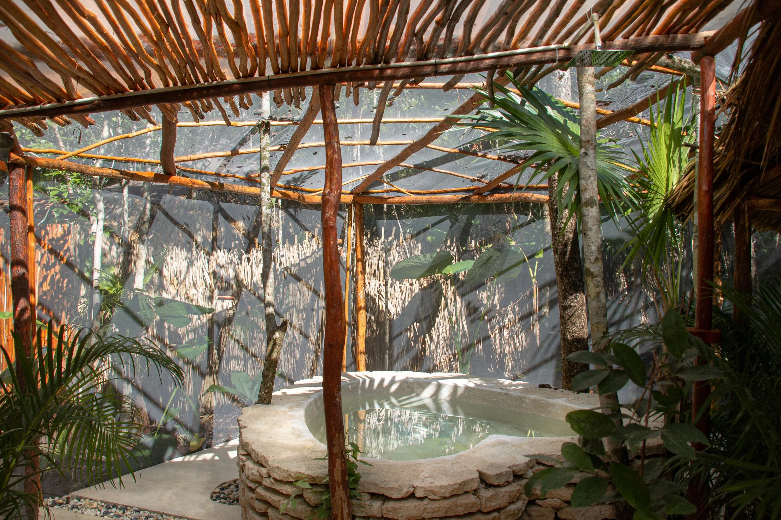 A rustic outdoor hot tub made of stacked stone, surrounded by tropical plants and under a bamboo and thatch roof structure with semi-transparent walls.