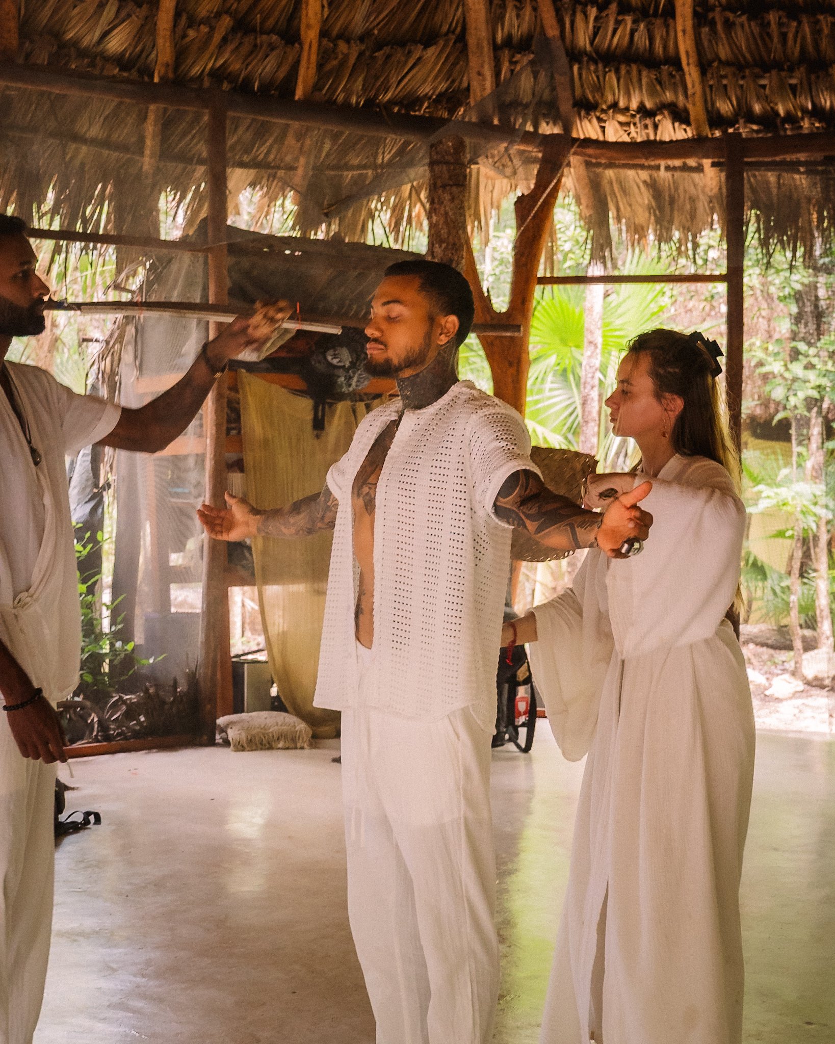 Man with tattoos standing with arms outstretched in a tropical outdoor hut, woman in white beside him, another man applying makeup or face paint, surrounded by lush green foliage.