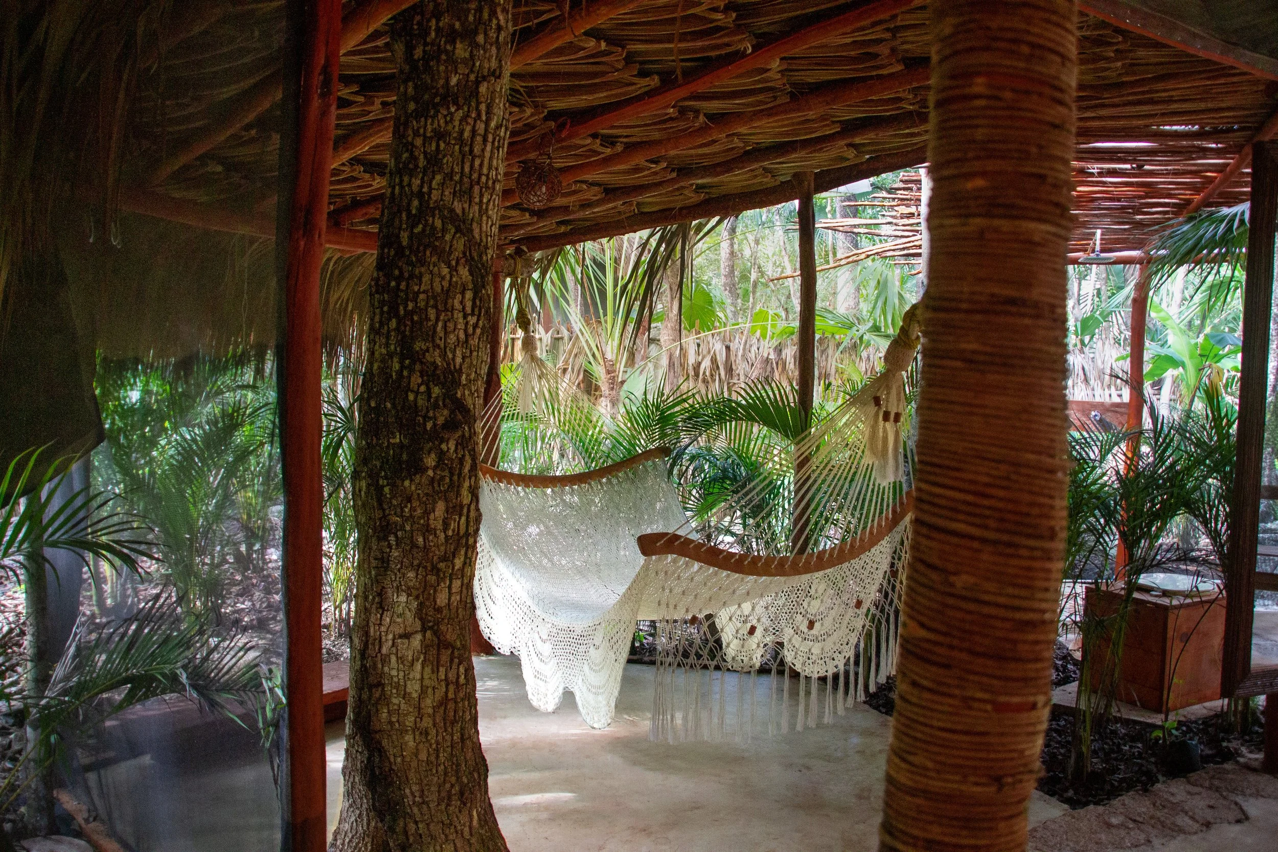 A hammock with a crochet cover is hanging outdoors among lush green tropical plants, wooden poles, and a thatched roof structure, with a trail and trees visible in the background.