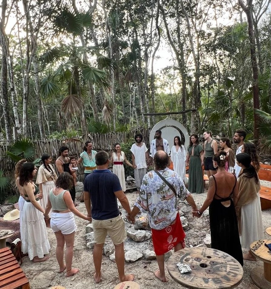 Group of people holding hands in a circle in a wooded outdoor setting, participating in a ceremony or ritual.