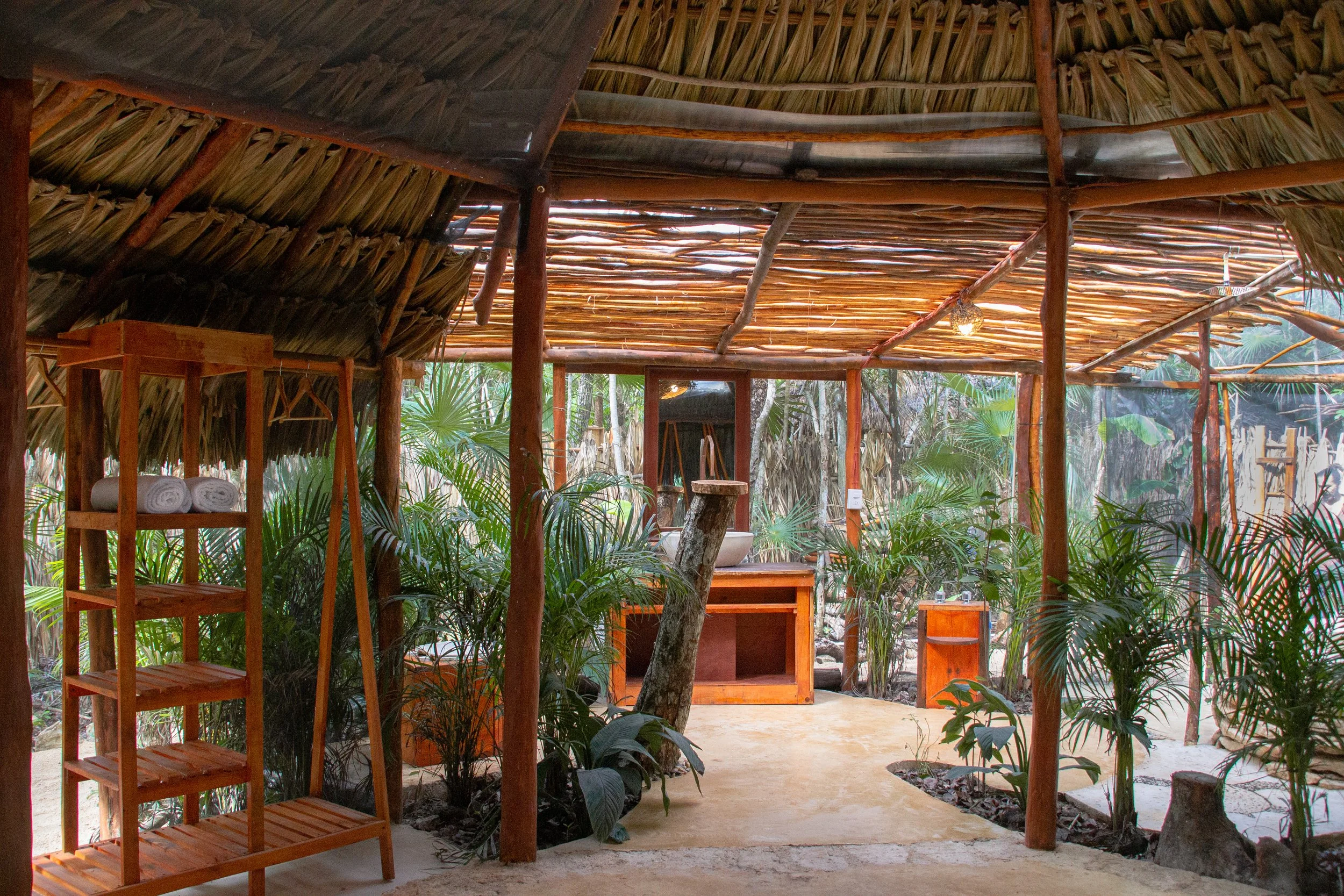 Interior of a rustic tropical outdoor shower area with a thatched roof, wooden shelves with towels, a mirror, and lush green plants surrounding the space.