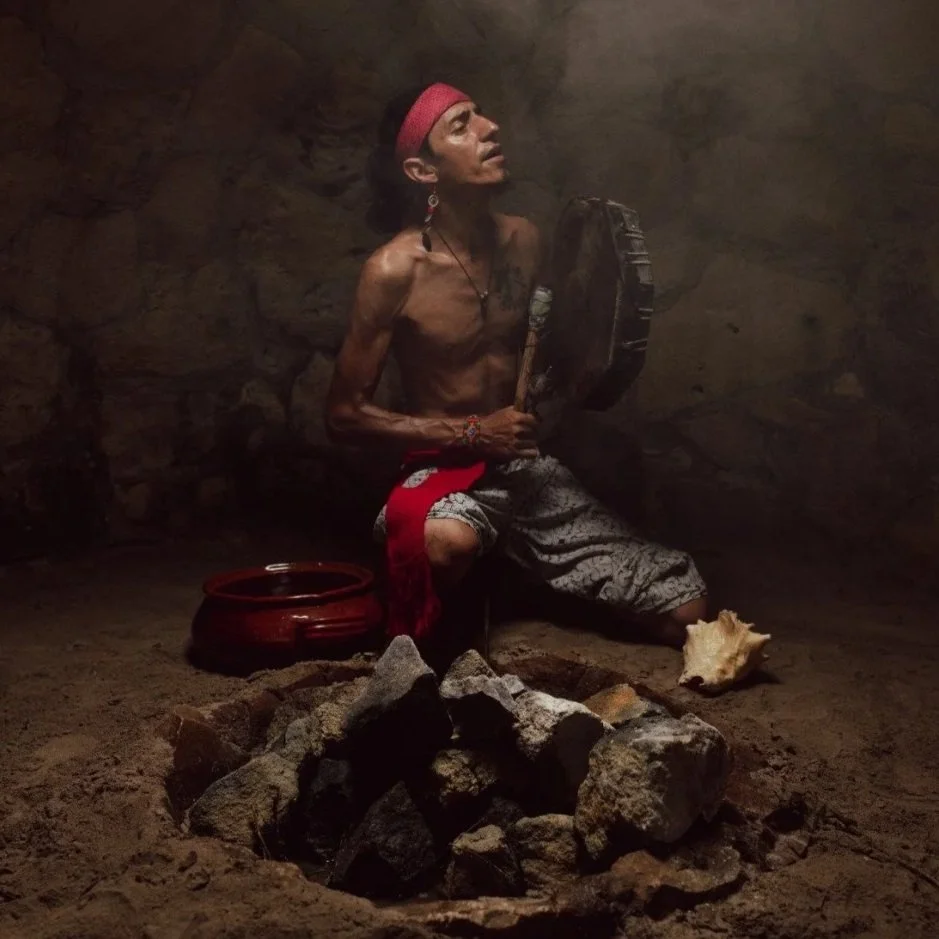 A shirtless man sitting on the ground inside a cave, holding a drum with a feather in his hand, surrounded by rocks and a large shell on the ground.