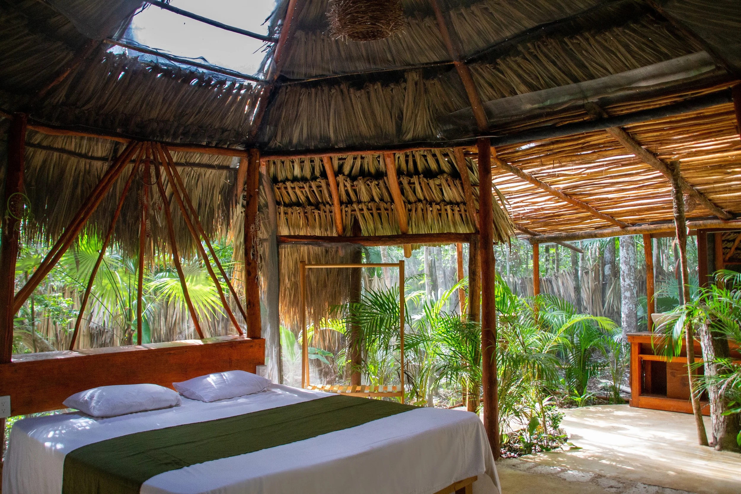 A bedroom in a rustic, open-air hut with a thatched roof, a bed with white linens and pillows, surrounded by lush greenery and tropical plants outside.