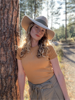 A young woman with long curly hair wearing a large sunhat, a yellow t-shirt, and gray pants, standing outdoors next to a tree in a forested area.