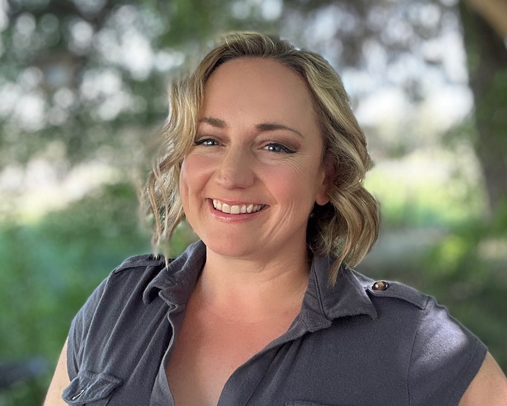 A woman smiling with blonde hair, wearing a black top, against a neutral background.